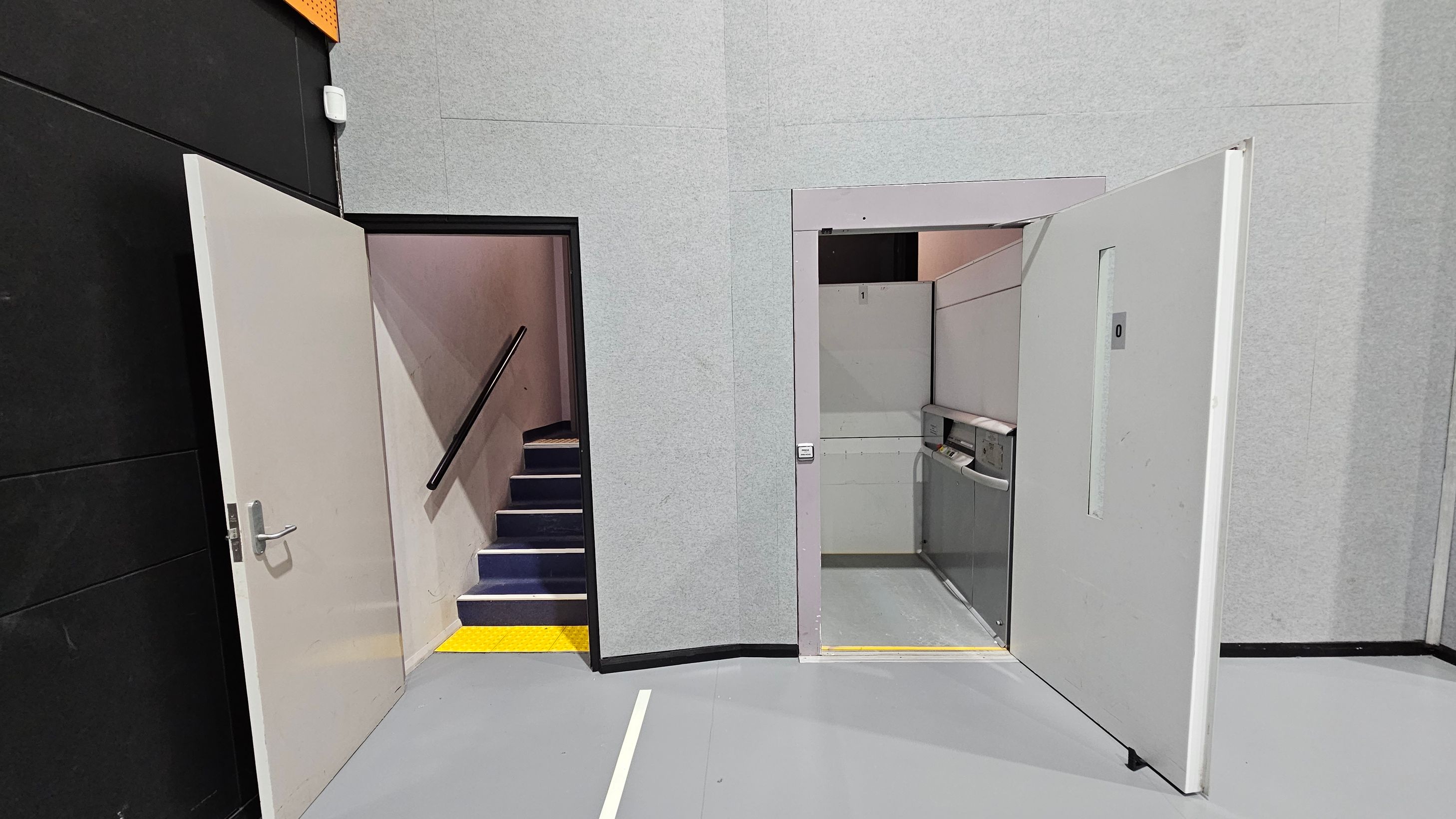 On the left, a door is open, revealing a staircase with a black handrail and blue steps, bordered by a yellow strip. On the right, another open door leads to a small lift with control panels visible. The walls are grey, and the floor is a smooth, light grey surface. The ceiling is not visible.