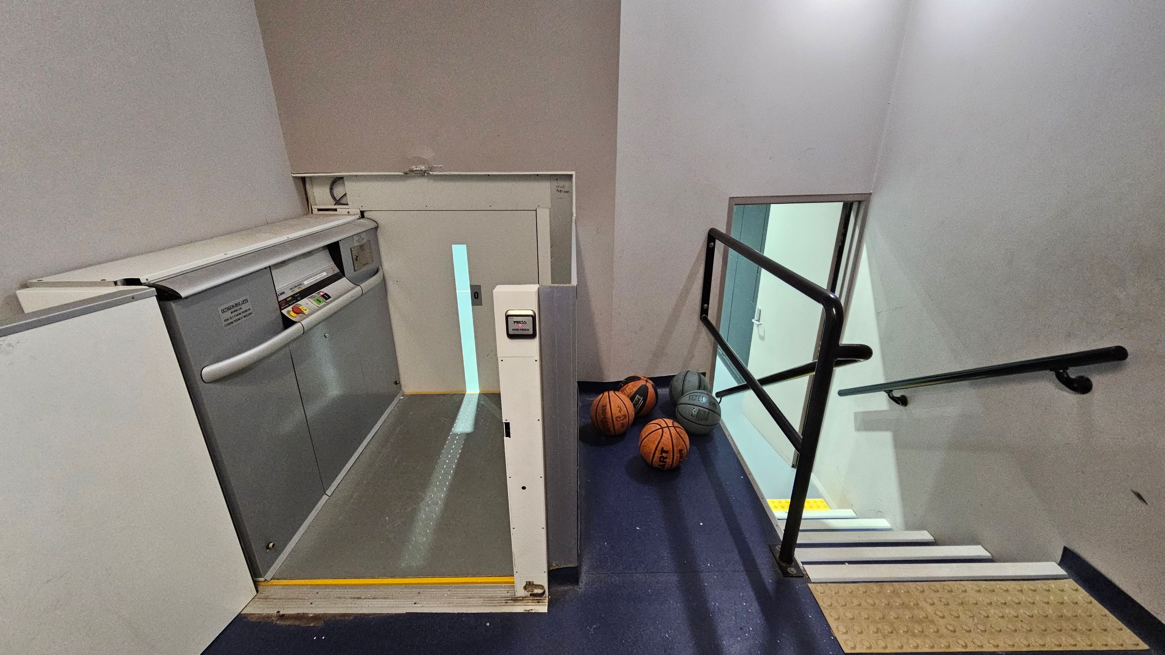 A stairwell with a platform lift on the left, featuring a control panel and safety rails. On the right, a set of stairs descends with black handrails. At the bottom of the stairs, several basketballs are scattered on the floor. The walls are light-coloured, and the floor is dark. The ceiling is not visible.