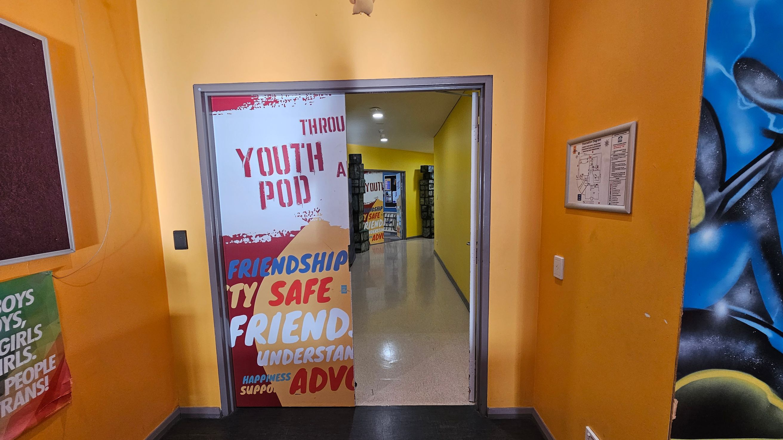 A hallway with bright yellow walls. On the left, a partially visible poster with colourful text, including "YOUTH POD" and words like "FRIENDSHIP" and "SAFE". A bulletin board is also on the left. On the right, a framed map or notice is mounted on the wall, and part of a blue and black mural is visible. The floor is shiny and light-coloured, leading to another room with similar decor. Ceiling lights illuminate the space.