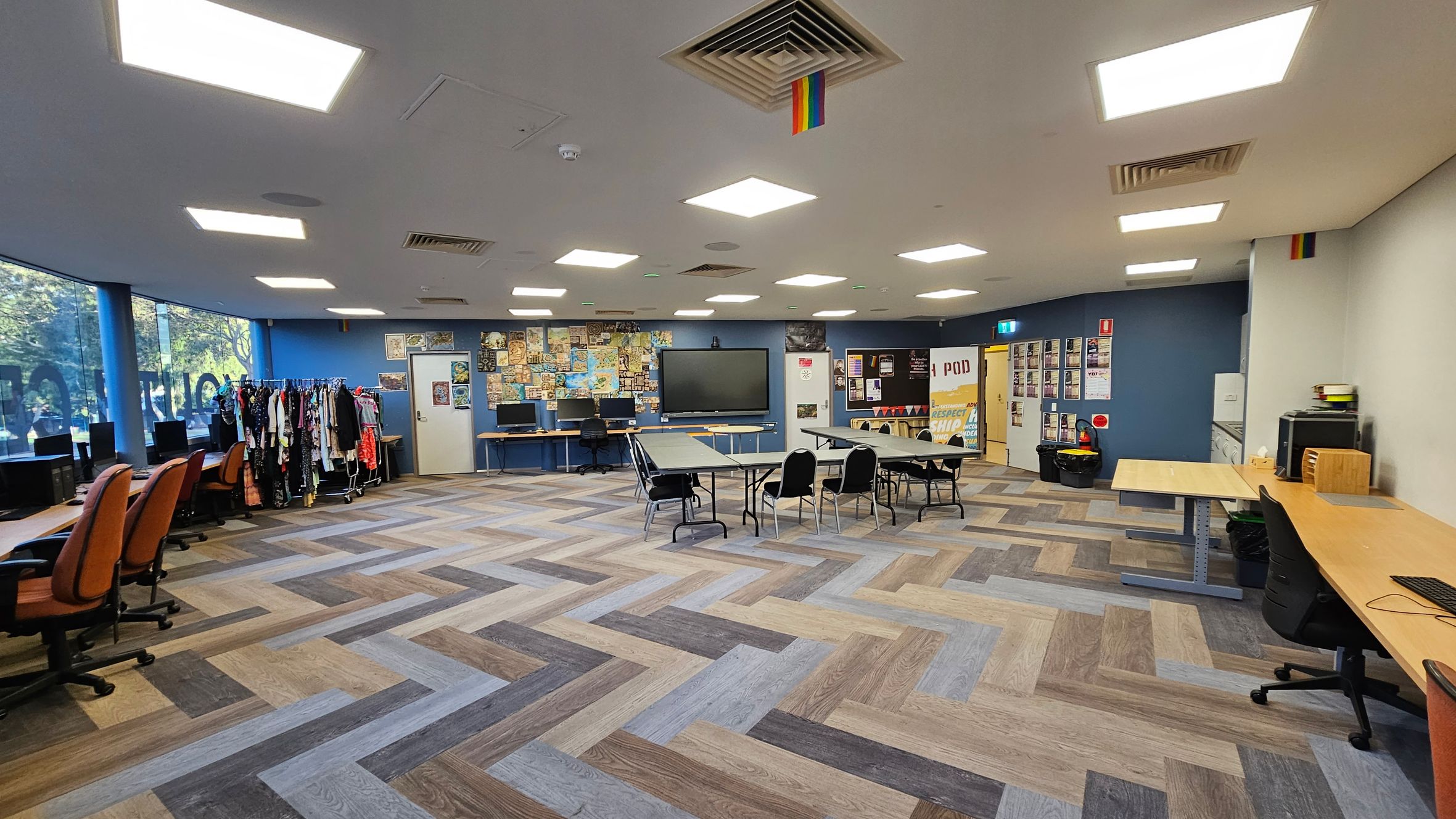 A spacious room with a herringbone-patterned floor in various shades of brown and grey. On the left, there are several orange office chairs in front of computer desks, with a clothing rack nearby. The centre features a large screen on the wall, surrounded by a collage of posters. In the middle, tables and chairs are arranged. On the right, more desks and chairs are visible. The ceiling has bright square lights and a rainbow flag. The walls are blue and white, with more posters and a door.