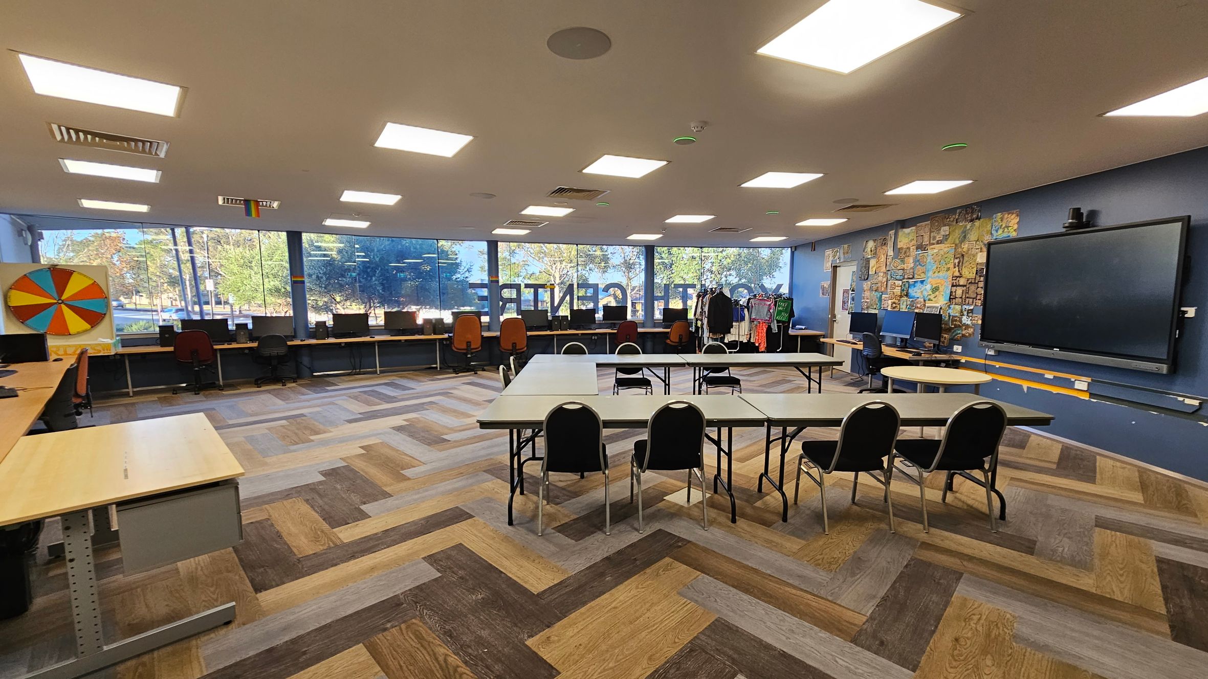 A spacious room with a herringbone-patterned wooden floor. On the left, a colourful wheel is mounted on the wall above a row of desks with computers and chairs. The back wall features large windows with a view of trees outside. In the centre, several tables are arranged in a U-shape with chairs around them. On the right, a large screen is mounted on a wall decorated with various posters and images. The ceiling has multiple rectangular lights.