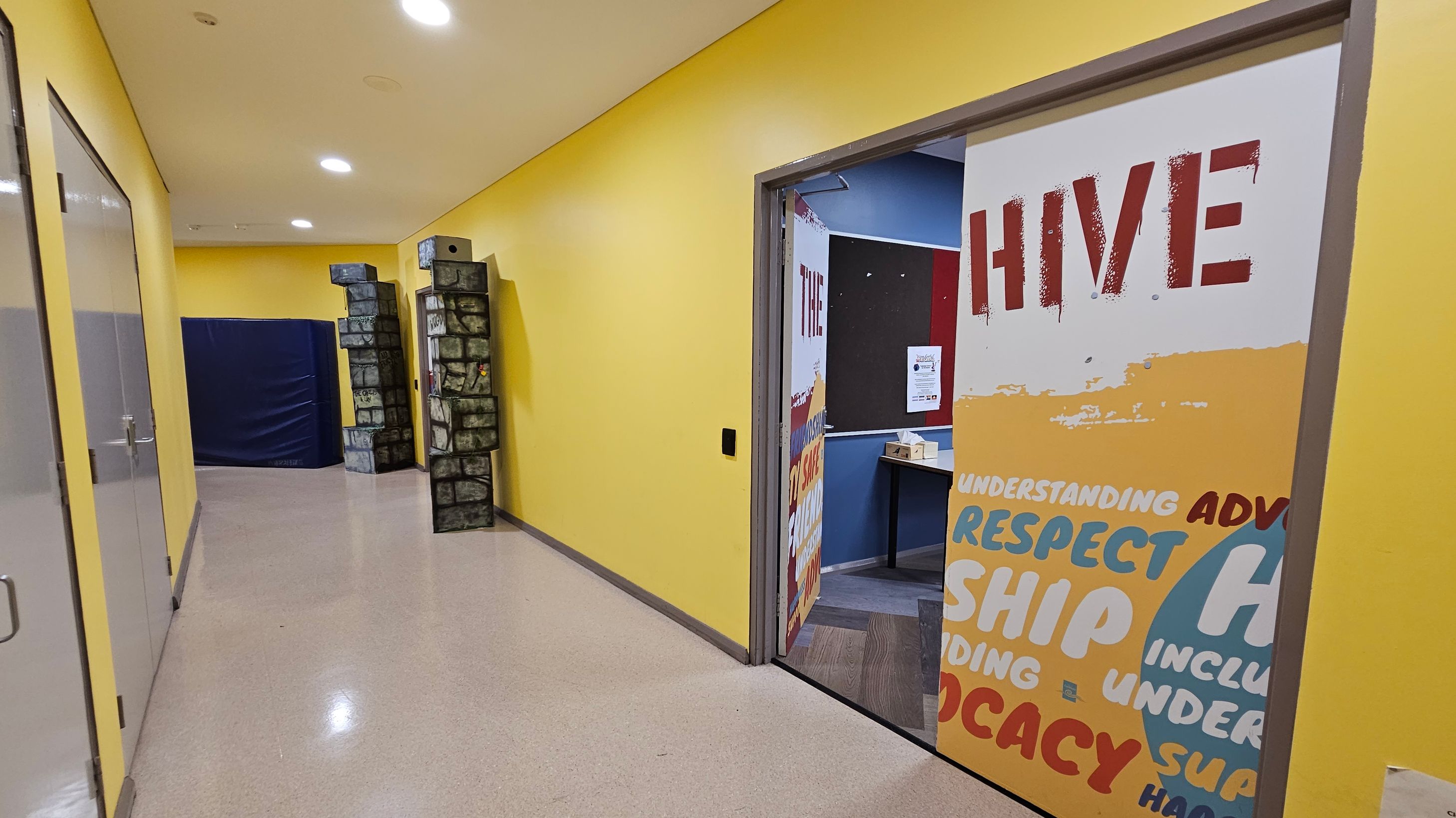 A hallway with yellow walls and a light-coloured floor. On the right, an open door with the word "HIVE" in large red letters and other words like "RESPECT" and "ADVOCACY" in various colours. Inside the room, a bulletin board and a table are visible. On the left, glass doors with metal handles. At the end of the hallway, there are two tall structures resembling stone pillars and a large blue padded mat. The ceiling has recessed lighting.