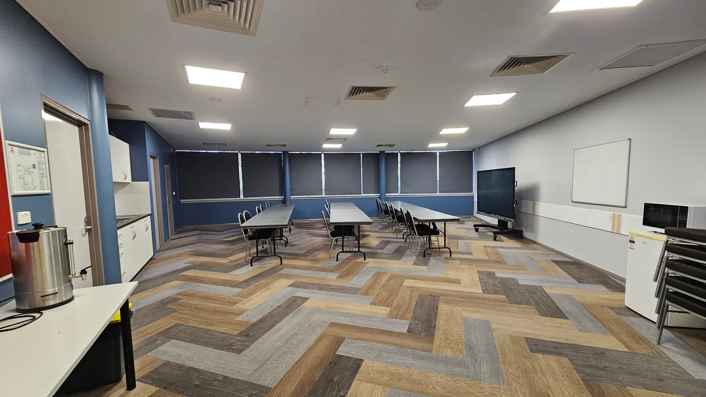 A room with a herringbone-patterned wooden floor and a white ceiling with recessed lights. On the left, there's a counter with a large metal urn and a door. Cabinets and a sink are along the left wall. In the centre, several tables with chairs are arranged in rows. The right side has a large screen on a stand, a whiteboard, and a small fridge with a microwave on top. The windows at the back are covered with dark blinds.