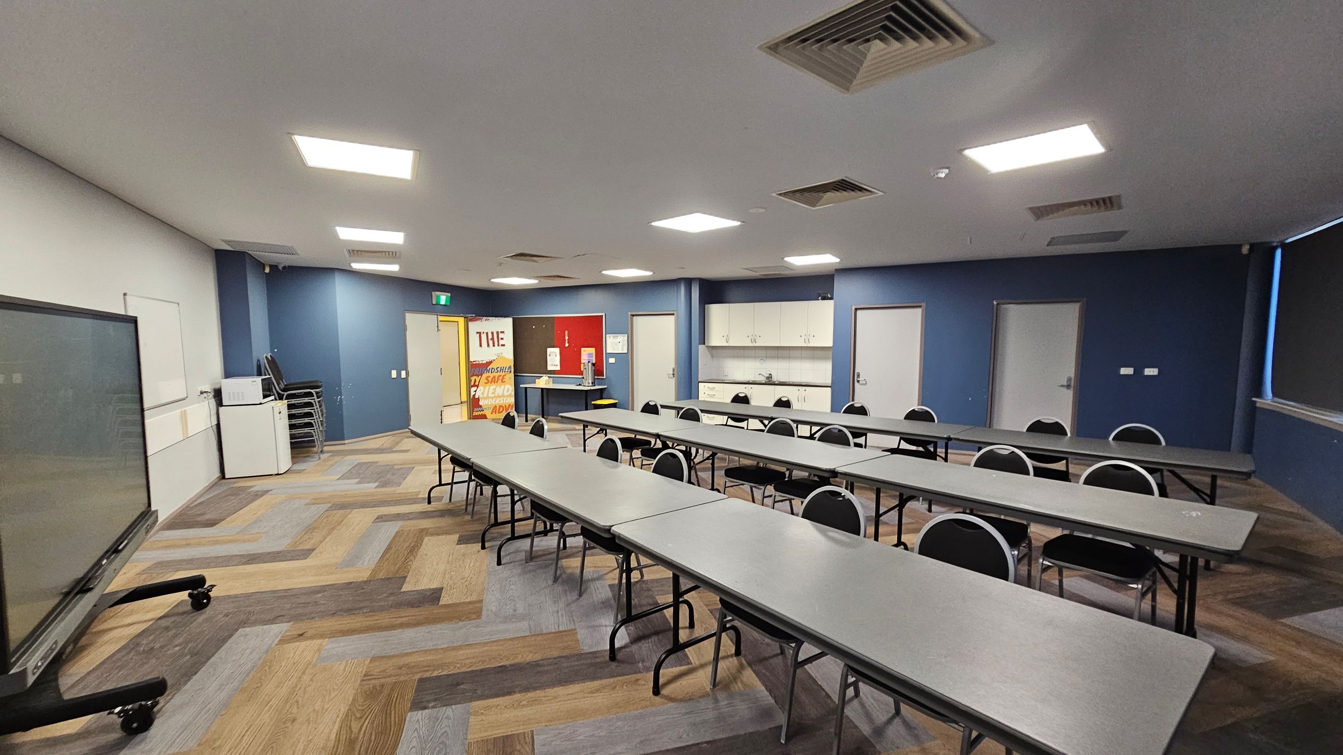 A classroom with a modern design. On the left, there's a large interactive screen on wheels and a whiteboard. The floor has a herringbone pattern with various shades of wood. In the centre, there are long tables with black chairs arranged in rows. The back wall is blue, featuring a door, cabinets, and a sink. On the right, a window with blinds is partially visible. The ceiling has recessed lighting and air vents. A colourful poster is near the back wall.