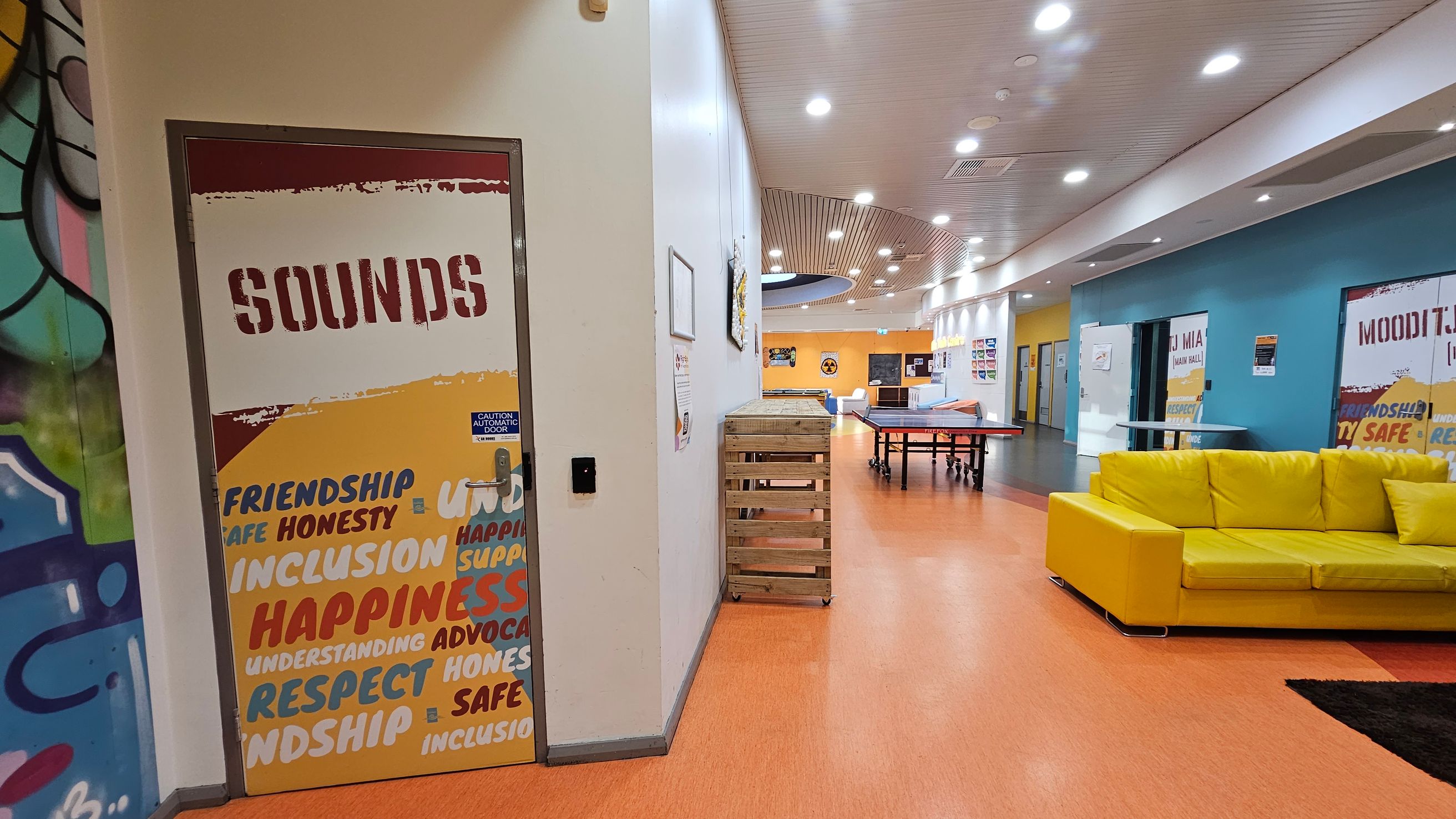 A brightly lit indoor space with an orange floor and a white ceiling featuring recessed lights. On the left, a door labelled "SOUNDS" is decorated with words like "FRIENDSHIP" and "HAPPINESS." Adjacent is a wall with colourful graffiti. In the centre, a wooden crate and a ping pong table are visible. On the right, a vibrant yellow sofa sits against a blue wall with more labelled doors. The corridor extends into the distance, with various posters and signs on the walls.