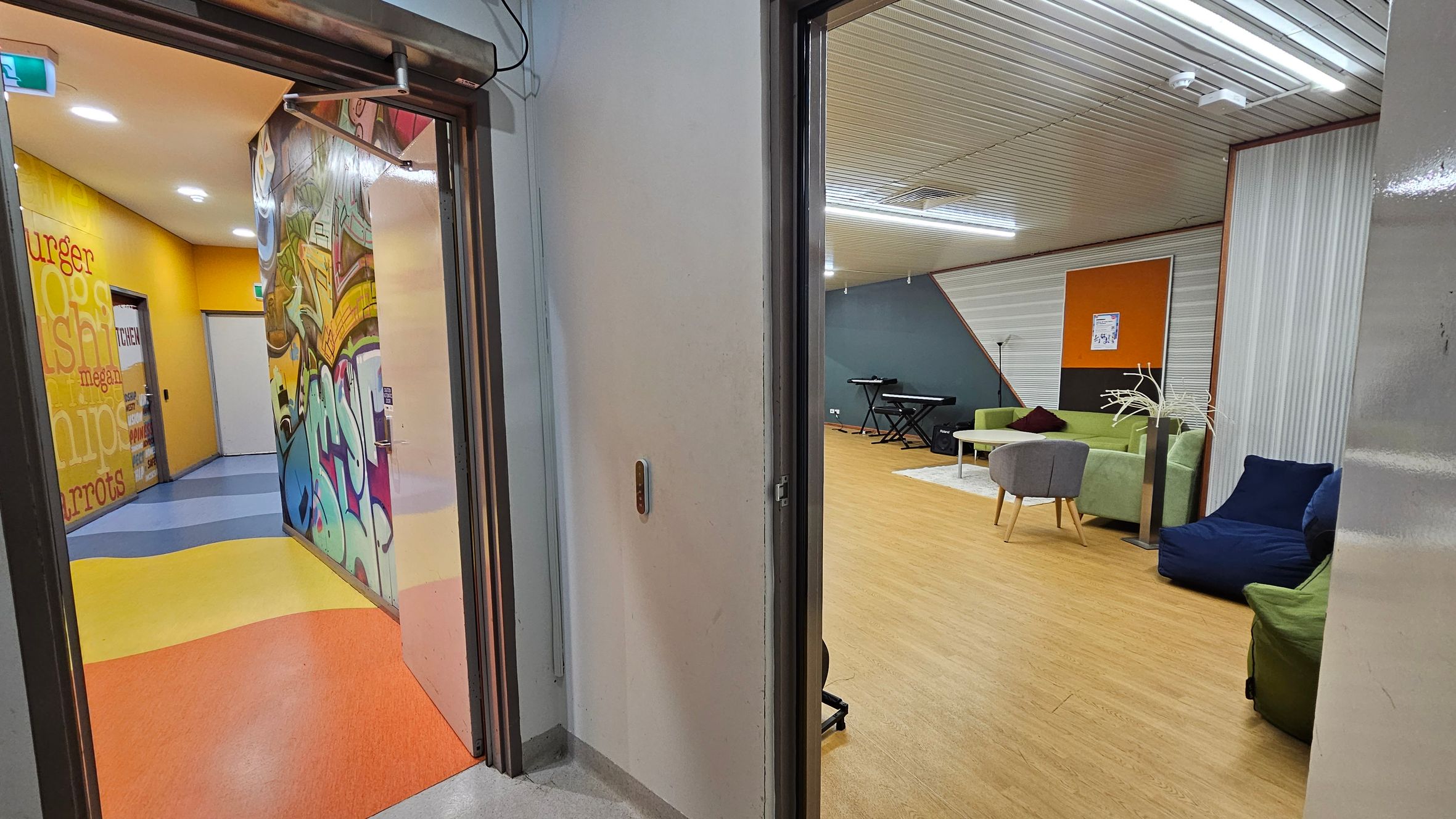 On the left, a hallway with vibrant graffiti art and a yellow wall featuring words like "burger" and "chips." The floor is a mix of blue, yellow, and orange. On the right, a room with a wooden floor, green and blue seating, a grey chair, and a keyboard against a blue wall. The ceiling is lined with white panels and fluorescent lights.