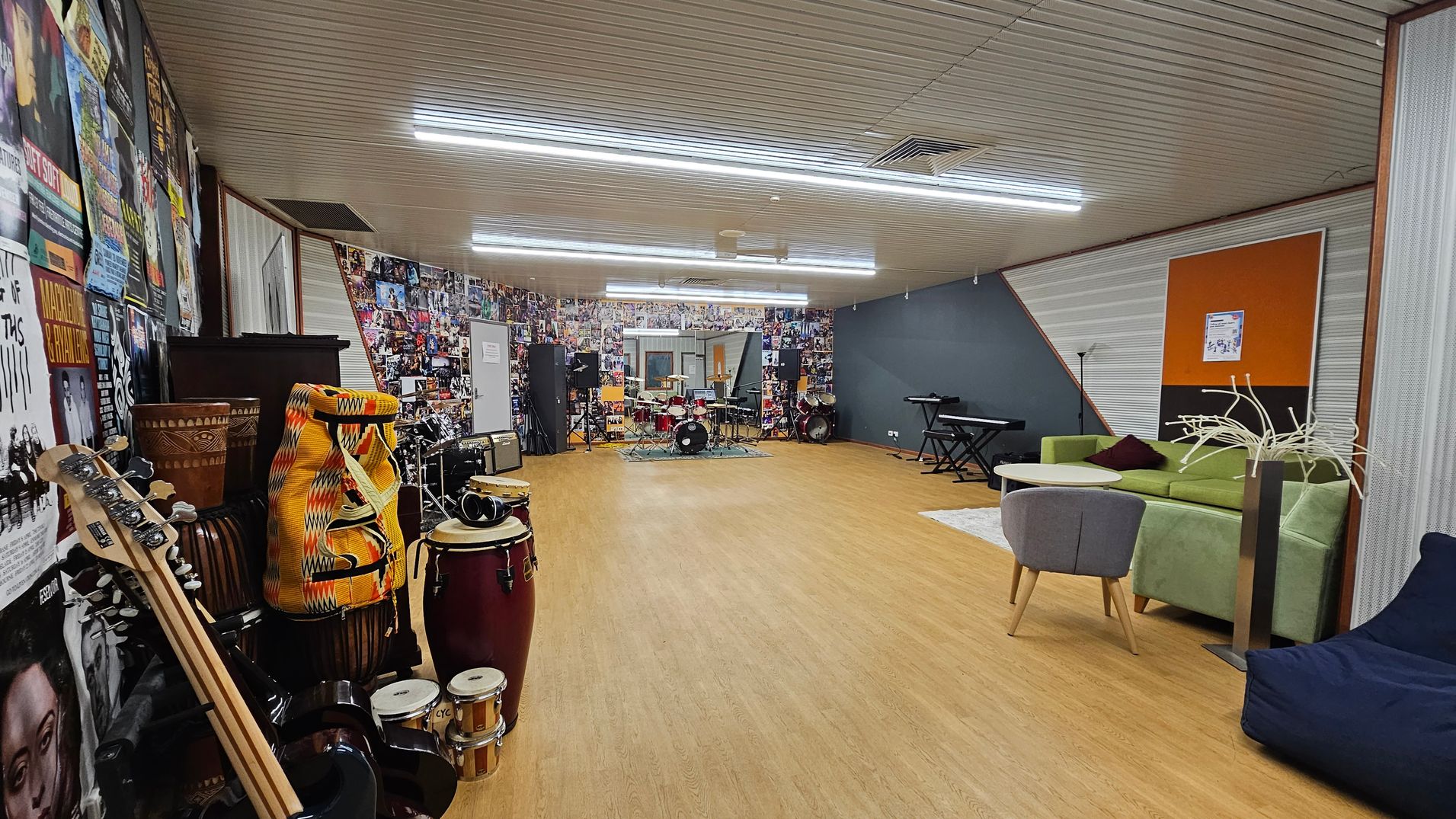 A spacious music room with a wooden floor and a ceiling with fluorescent lights. On the left, there are various musical instruments, including guitars, drums, and percussion instruments, against a wall covered in posters. In the centre, a drum set is visible. On the right, there is a green sofa, a small round table, a grey chair, and a blue bean bag. The back wall is decorated with numerous photos, and a keyboard is positioned nearby.