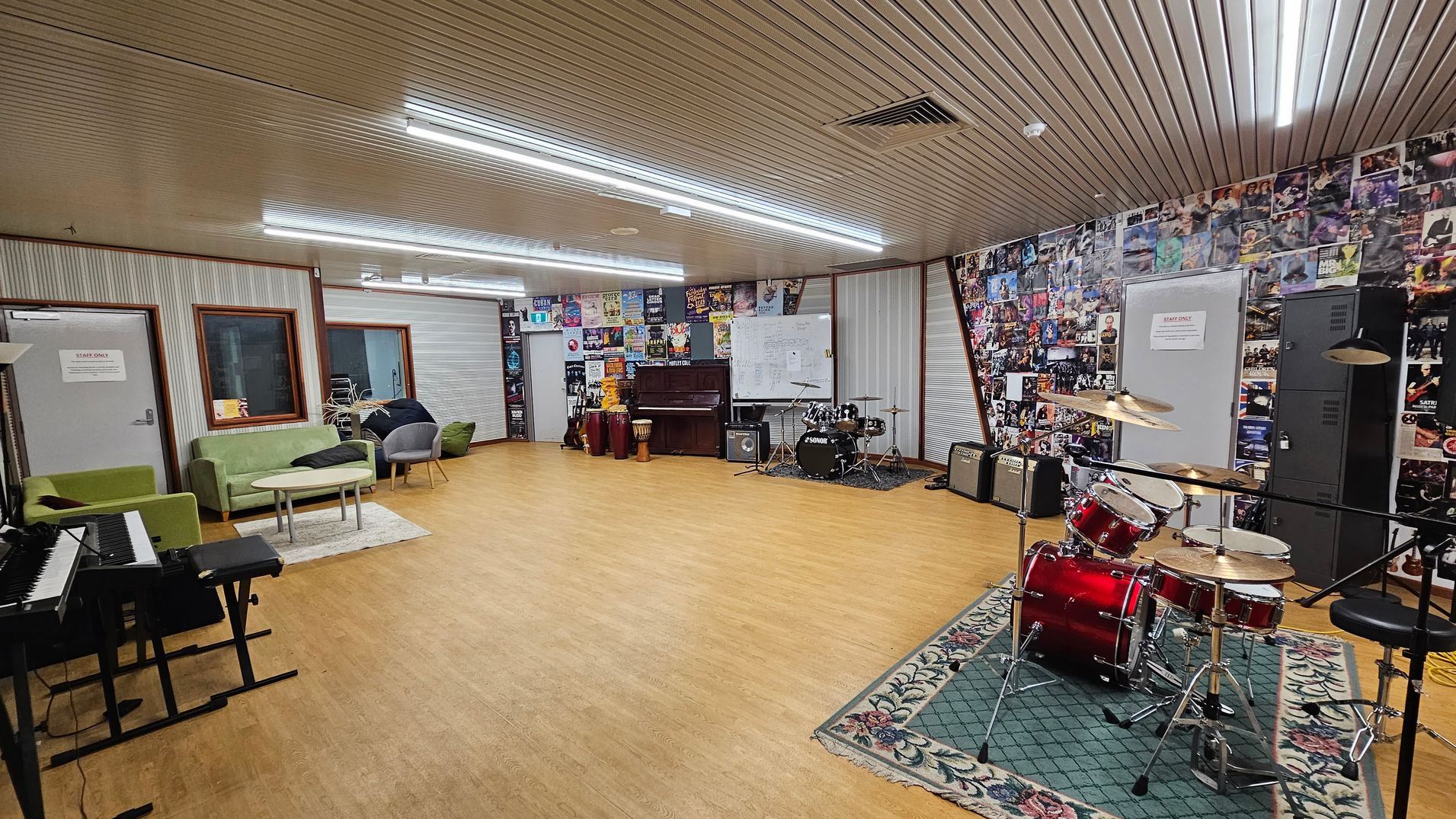 A music room with a wooden floor and a ceiling with fluorescent lights. On the right, a red drum set is placed on a patterned green rug. The back wall is covered with posters and features a piano, conga drums, and amplifiers. A whiteboard is also visible. On the left, there are green sofas and grey chairs around a small table, with a keyboard nearby. The walls have a mix of corrugated and poster-covered surfaces.