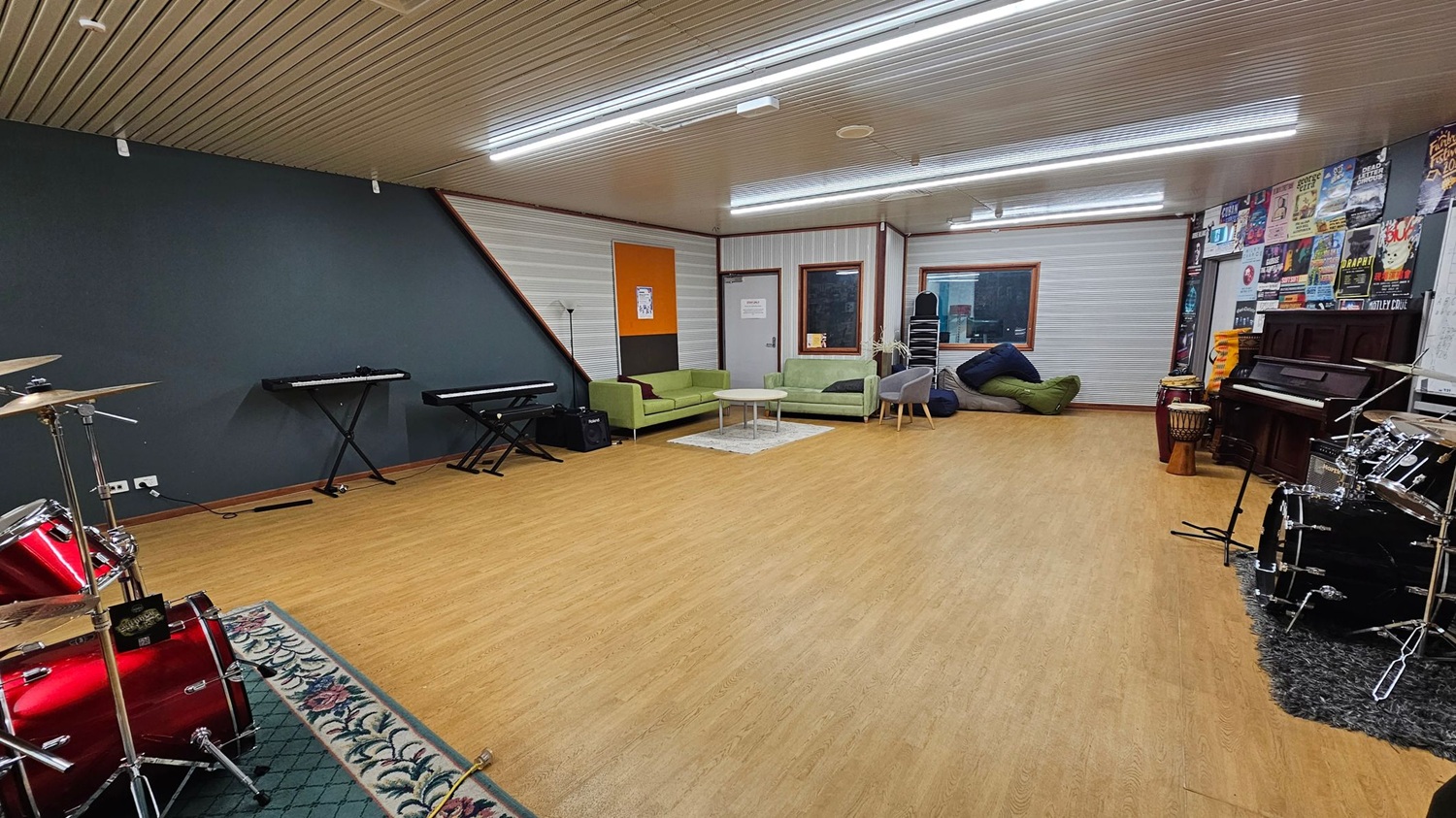 A spacious music room with a wooden floor and a ceiling with fluorescent lights. On the left, there are two electronic keyboards against a dark wall, and a red drum set on a patterned rug. In the centre, green sofas and a small table are placed on a light rug. The right side features a wall with posters, a piano, and additional drums. A window and a door are visible in the background, along with stacked chairs and bean bags.