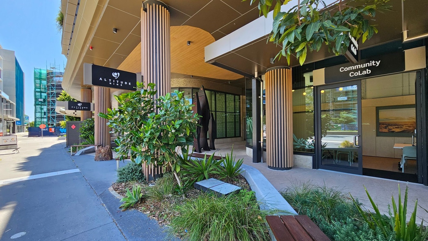 A modern building entrance with a sign reading "Altitude Nine" on the left and "Community CoLab" on the right. The area features tall, cylindrical columns and lush greenery, including shrubs and small trees. The pavement is clean and grey, with a wooden bench nearby. Large glass windows and doors are present, reflecting the surrounding urban environment. In the background, a building under construction is visible, covered in scaffolding and green netting.