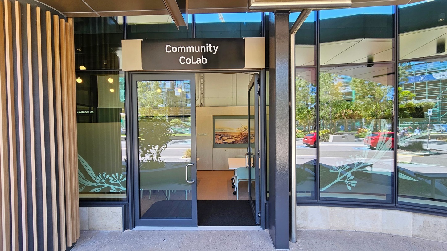 Entrance to a building with a sign reading "Community CoLab" above the door. The door is glass with metal handles, and the interior shows a table and chairs. The left side has vertical wooden slats, while the right side features large glass windows reflecting trees and parked cars outside. Inside, a framed picture is visible on the wall. The floor is concrete, and the ceiling has a dark finish with visible beams.