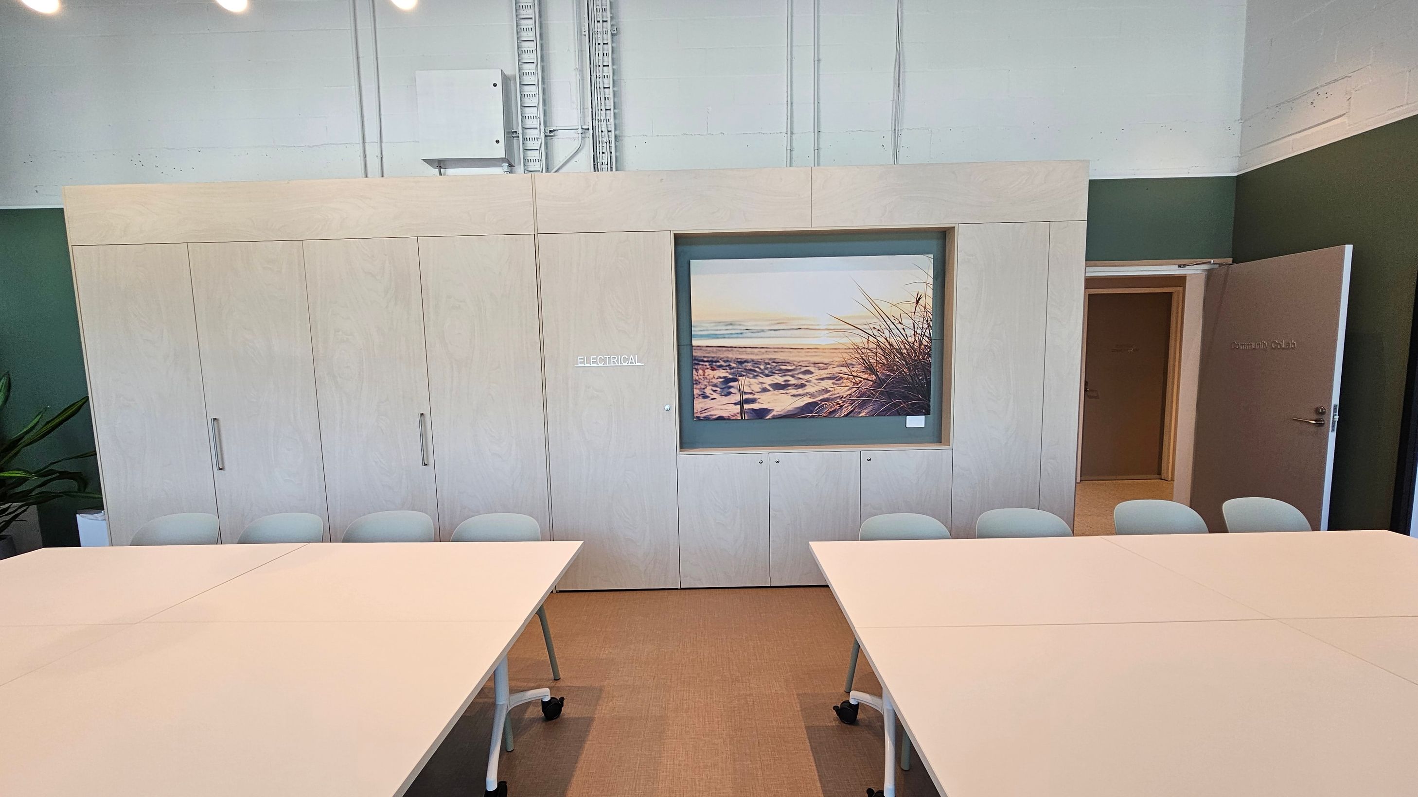 Entrance to a building with a sign reading "Community CoLab" above the door. The door is glass with metal handles, and the interior shows a table and chairs. The left side has vertical wooden slats, while the right side features large glass windows reflecting trees and parked cars outside. Inside, a framed picture is visible on the wall. The floor is concrete outside and appears to be carpeted inside.