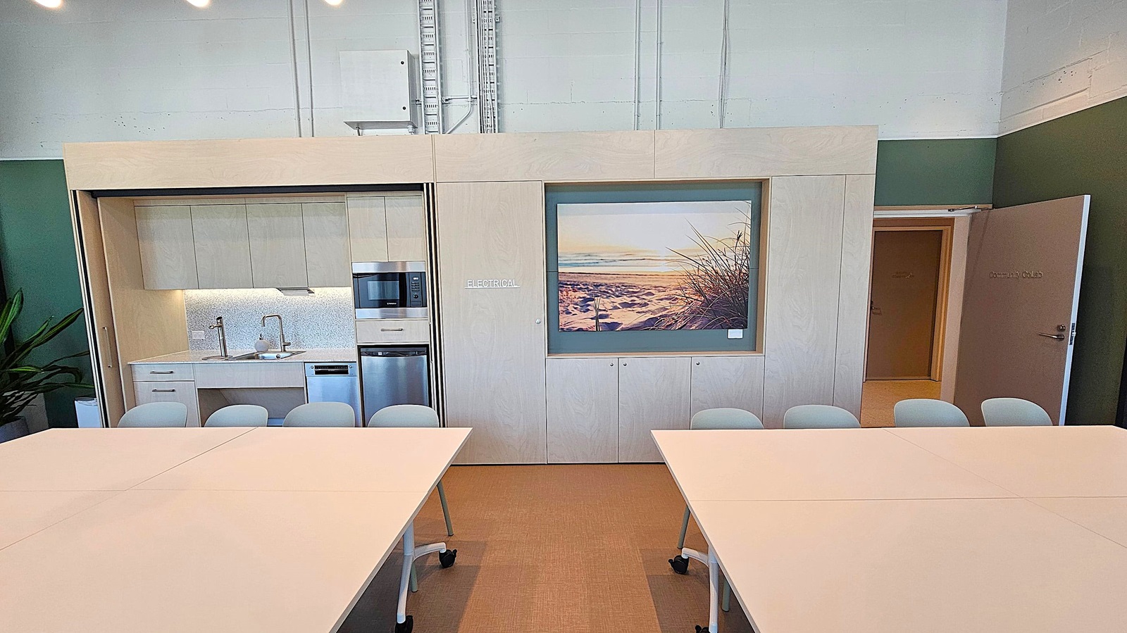 A room with light-coloured walls and a high ceiling. In the centre, there is a large cabinet with multiple doors, labelled "ELECTRICAL," and a framed picture of a beach scene. Below the picture, there are several light grey chairs. On the right, an open door leads to another room. In the foreground, two white tables on wheels are positioned with a gap between them. The floor is carpeted in a light brown shade.