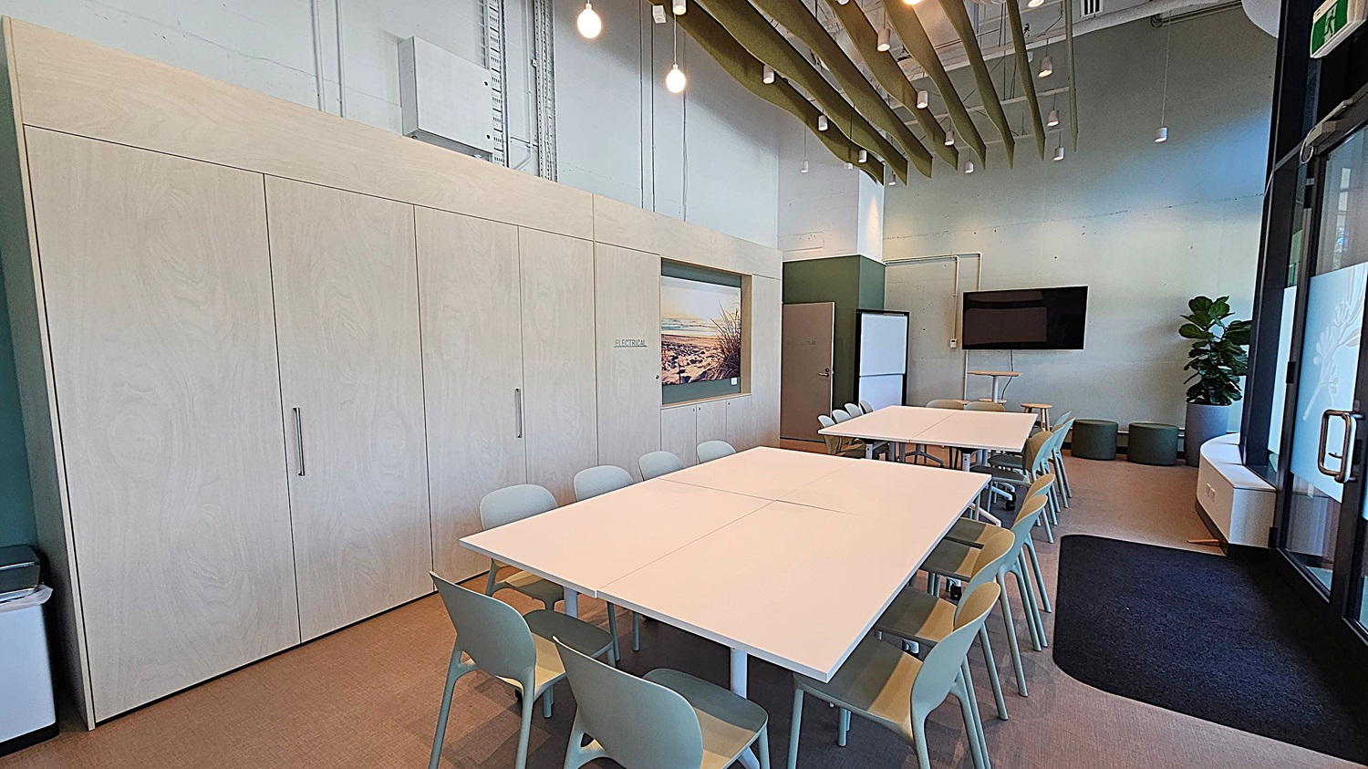 A modern office space with a kitchenette on the left, featuring white cabinets, a sink, and built-in appliances. In the centre, there's a large framed photograph of a beach scene. To the right, an open door leads to another room. In the foreground, there are white tables with chairs on wheels. The walls are a mix of white and green, and the ceiling is high with visible pipes and lighting fixtures.