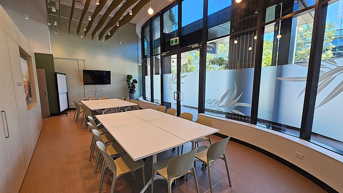 A modern meeting room with a large rectangular table in the centre surrounded by beige chairs. On the left, there is a wall with a door and a mounted flat-screen TV. The right side features large windows with frosted glass designs, allowing natural light to enter. A potted plant is near the corner. The ceiling has hanging lights and decorative wooden slats. The floor is carpeted in a light brown tone.