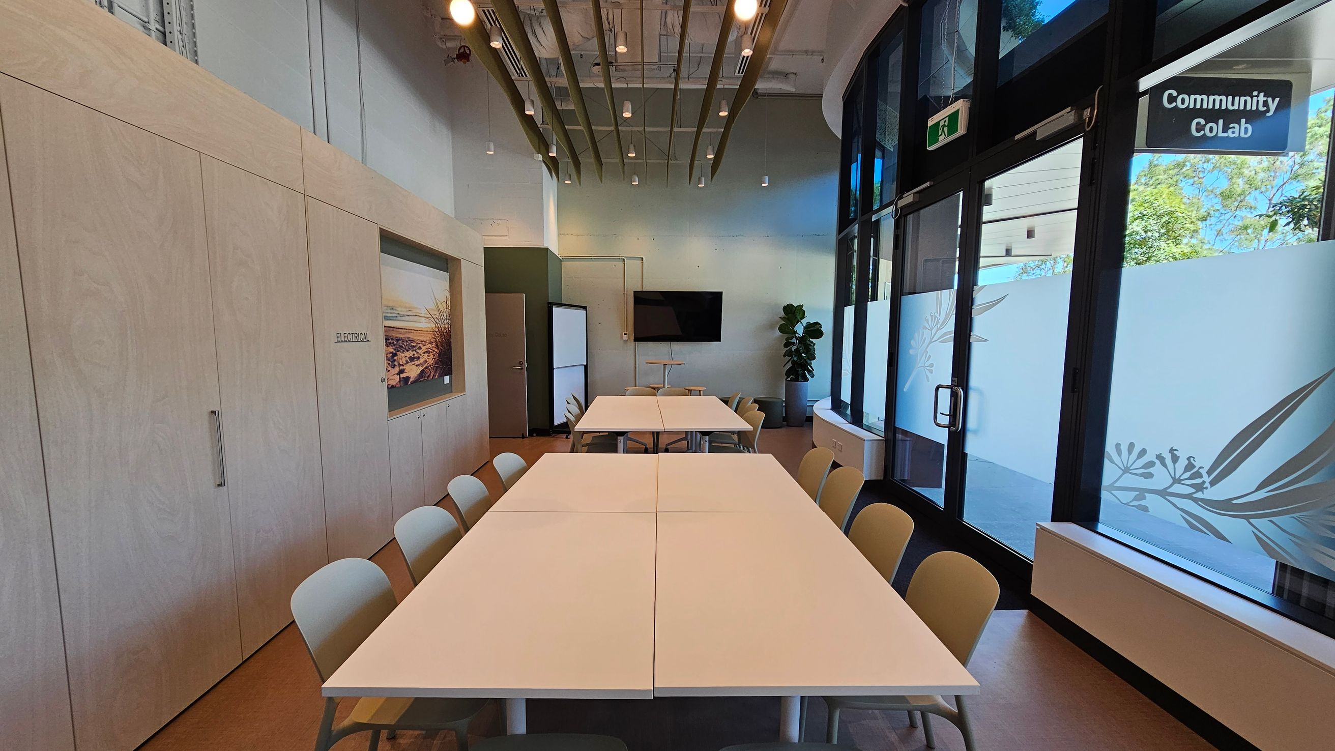 A modern meeting room with a long table in the centre surrounded by chairs. On the left, there are light wooden cabinets with a landscape picture. The right side features large windows with frosted glass and a "Community CoLab" sign. At the back, a TV is mounted on the wall, and a potted plant is nearby. The ceiling has exposed beams and hanging lights. The floor is a light brown colour.