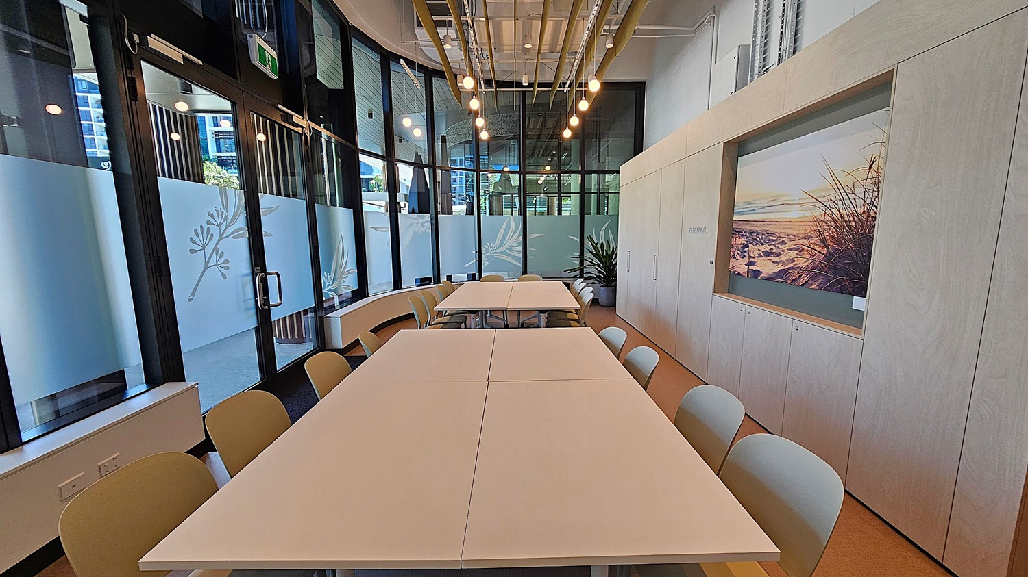 A modern meeting room with a long table in the centre surrounded by chairs. On the left, there are light wooden cabinets with a landscape picture. The right side features large windows with frosted glass and a "Community CoLab" sign. At the back, a TV is mounted on the wall, and a potted plant is nearby. The ceiling has exposed beams and hanging lights. The floor is a light brown colour.