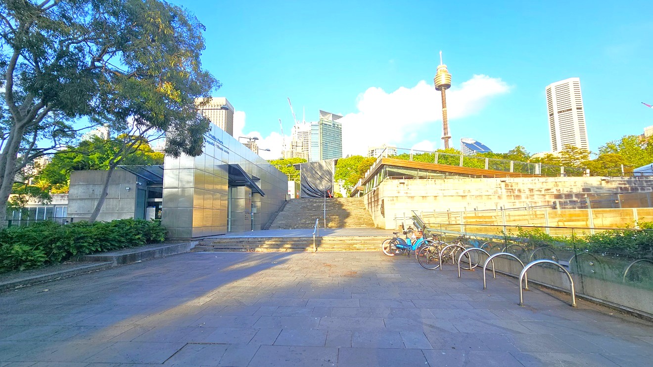 To the left is the facility building with silver walls. On the right, a bike rack holds a couple of bikes. In the middle, there are steps with a single handrail, leading to another set of steps for the upper level. Landscape ornamental plants and trees fill the plant box on the left. Tall trees and commercial buildings, including the Sydney Eye Tower, are visible.