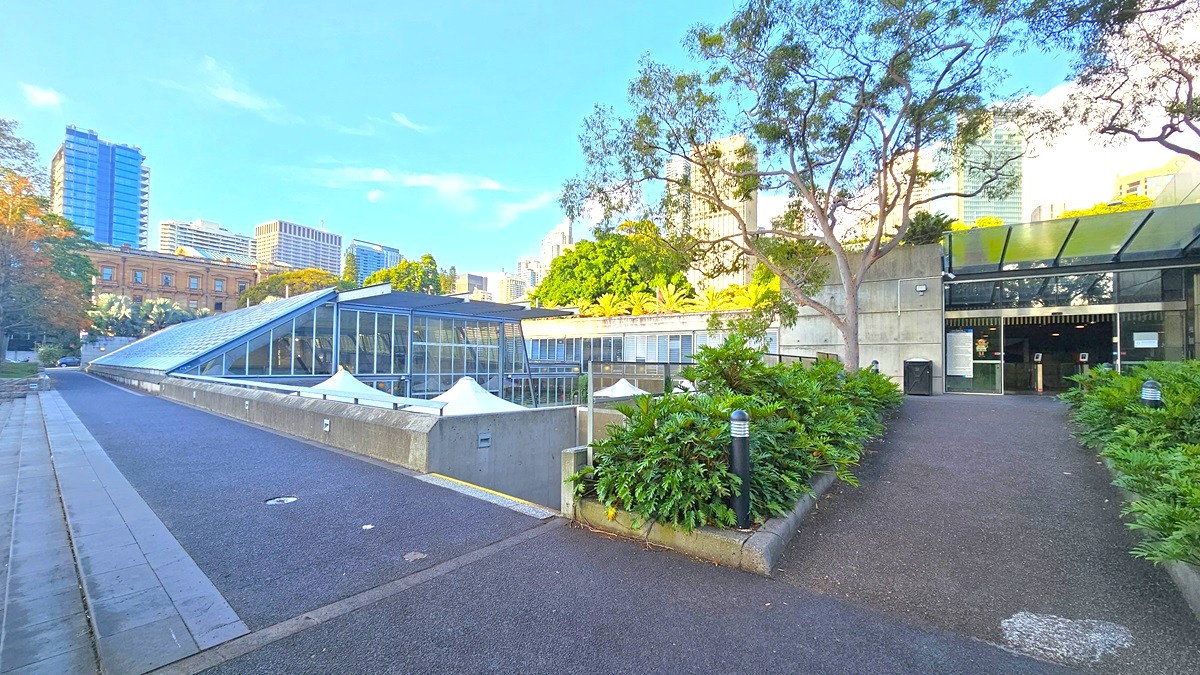 To the left, steps and a bitumen path extend near the building's rooftop. In the middle, the top portion of umbrella shades is visible. To the right, the bitumen path leads towards the entrance door. Two garden lamps are positioned among ornamental plants. The building's entrance features an extended glass roofing. Tall buildings and trees surround the area.