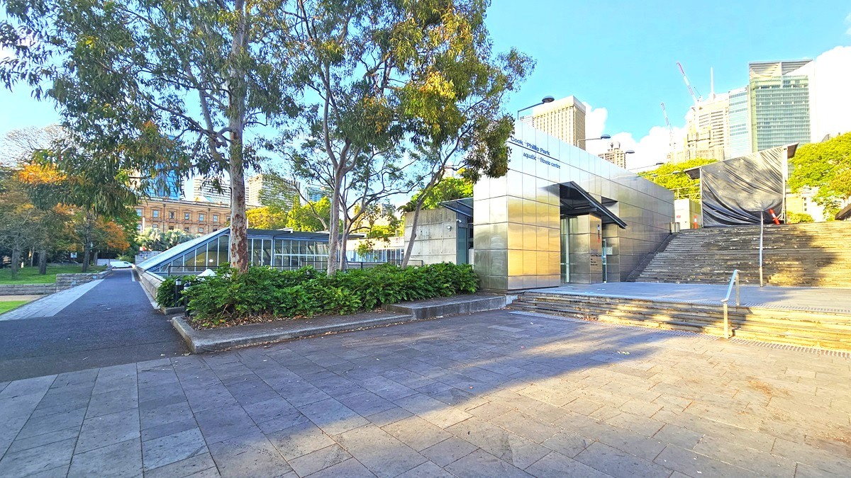 In the middle is the facility building with silver walls. On the right, there are steps with a single handrail, leading to another set of steps for the upper level. On top of the upper stairs is a black shade cloth covering a section. Landscape ornamental plants and trees fill the plant box on the left. Tall trees and commercial buildings, including the Sydney Eye Tower, are visible. A bitumen path leads to the left.