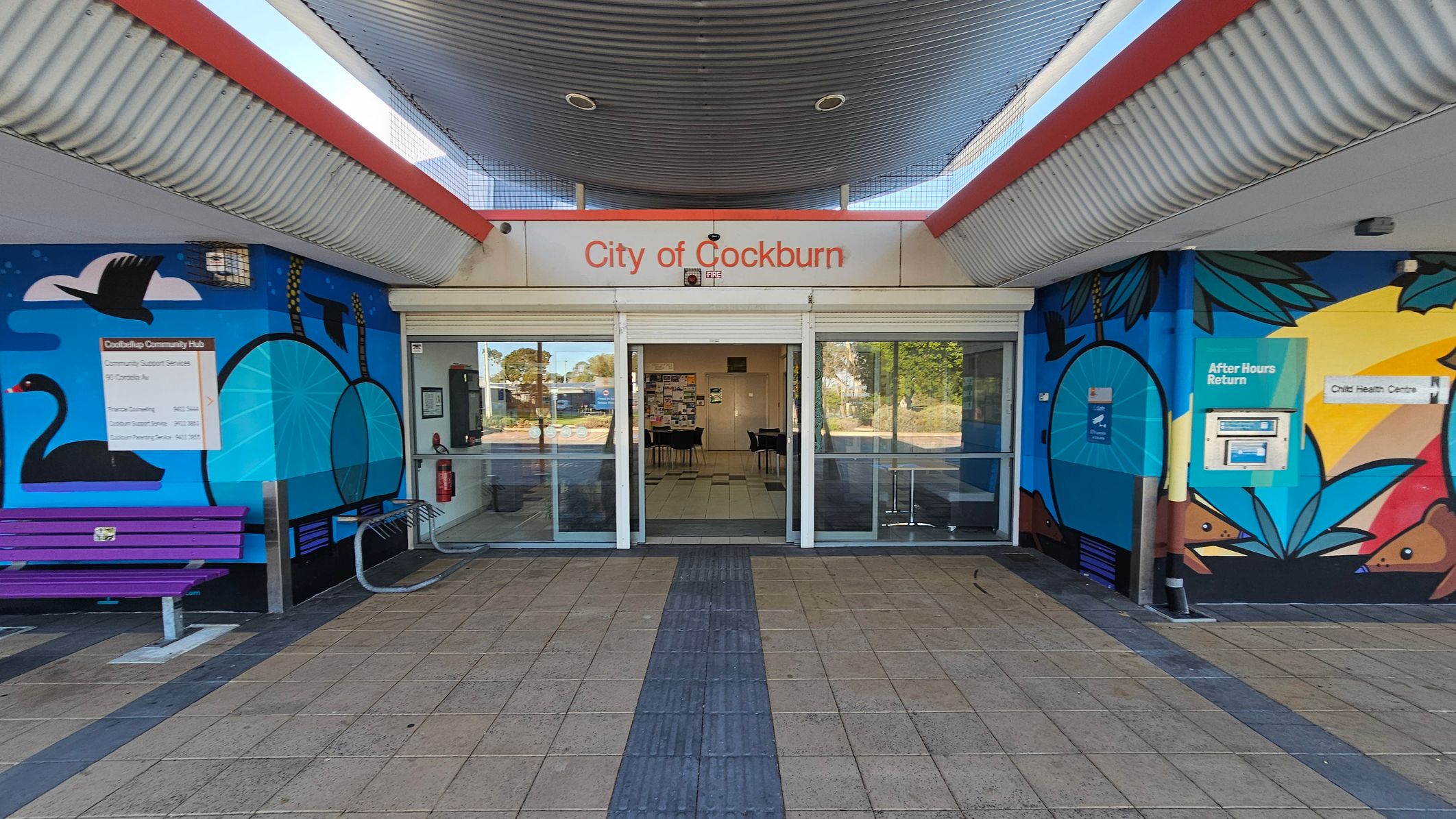 Entrance to a building with a sign reading "City of Cockburn" above glass sliding doors. The walls on either side feature colourful murals with abstract designs and images of swans. A purple bench is on the left, and an "After Hours Return" box is on the right. The floor is tiled with a central path leading to the entrance. The ceiling is corrugated metal with red accents. Inside, tables and chairs are visible through the glass doors.