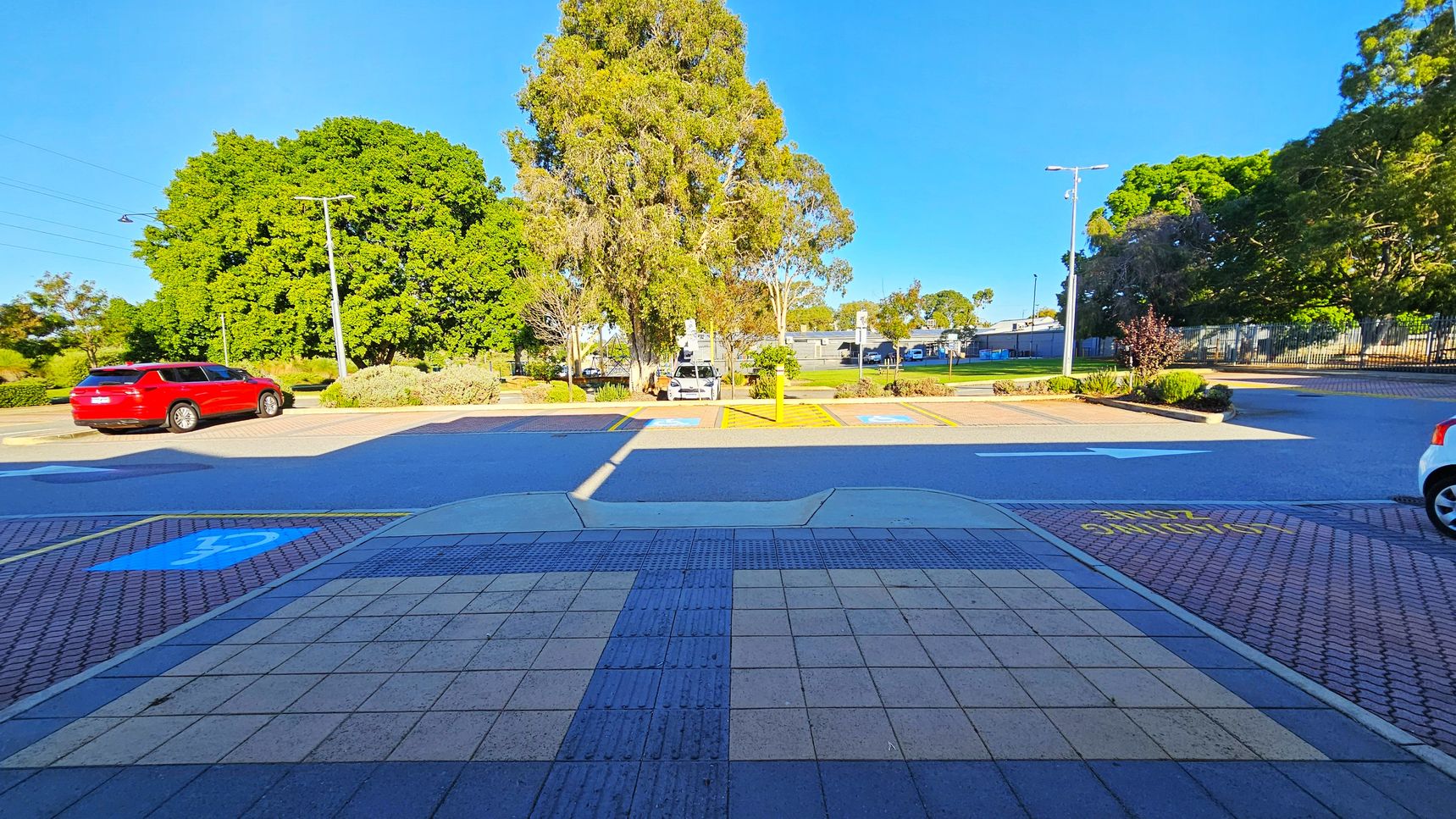 A parking area with a red car on the left and a white car partially visible on the right. In the centre, there are parking spaces marked with blue and yellow lines, indicating disabled parking. Large green trees are in the background, with a few streetlights and a building partially visible. The foreground has a tiled pavement with a textured strip, and the words "SLOW" painted in yellow on the ground.