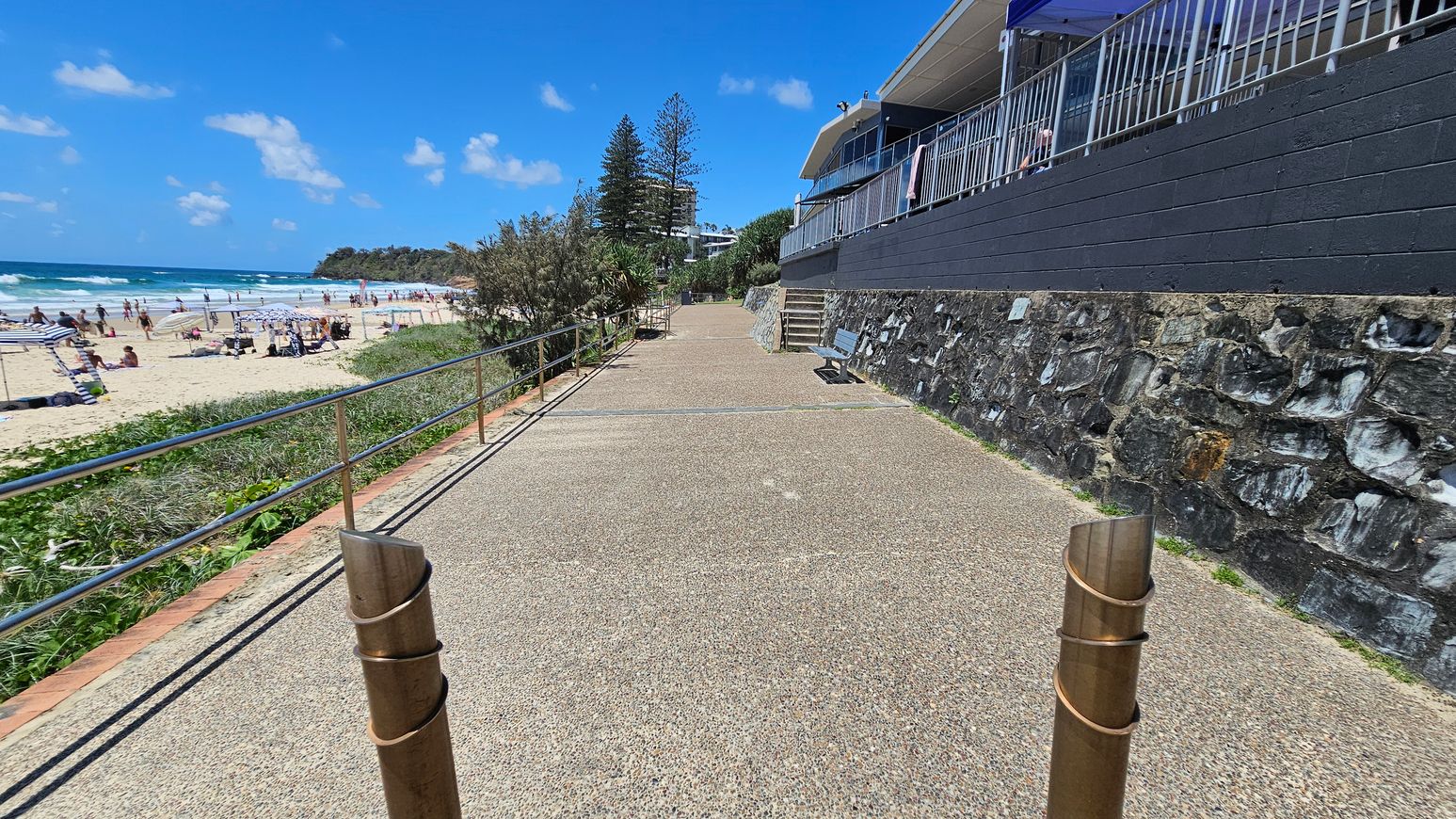 A coastal walkway with a textured path runs alongside a stone wall on the right, topped with a building featuring a metal railing. On the left, a sandy beach is visible with people sunbathing and swimming. The beach is bordered by greenery and a metal railing. In the distance, trees and a rocky outcrop are visible. The sky is clear with a few clouds.