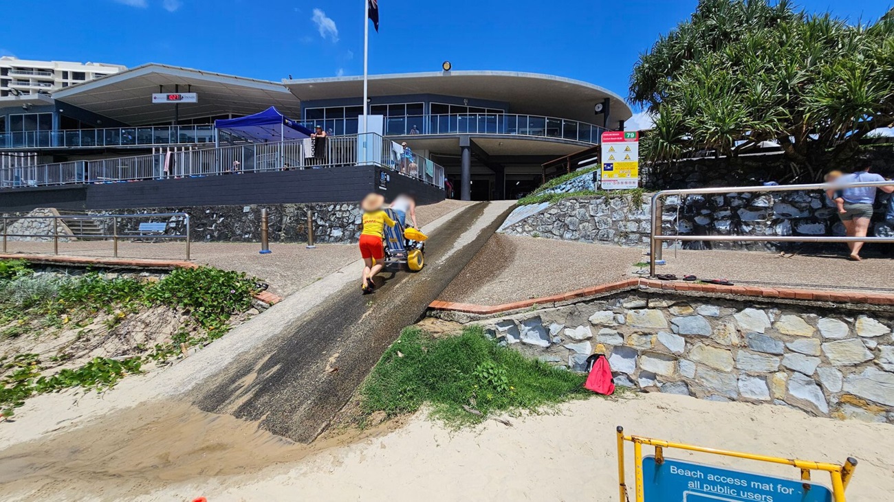 A sandy beach area with a ramp leading up to a building. Two people are pushing a wheelchair up the ramp. The building has a modern design with large windows and a flat roof. A stone wall lines the ramp, and there are signs and railings along the path. On the right, a person is walking up a separate path. Greenery is visible on the left side near the sand. A yellow sign at the bottom of the image indicates beach access.