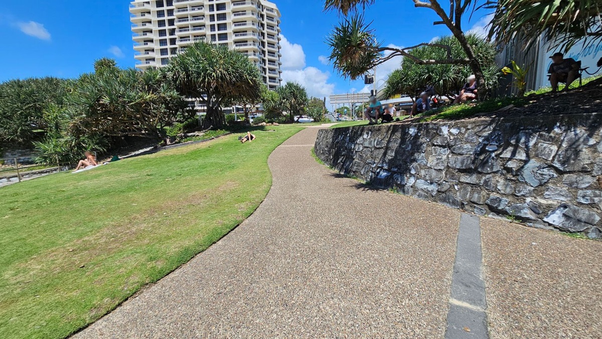 A curved pathway runs through a park with green grass on the left and a stone wall on the right. People are sitting on the grass and along the wall. In the background, there are tall trees and a high-rise building.