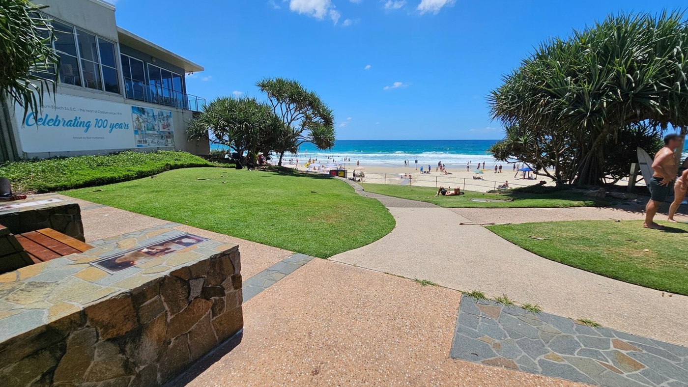 A coastal scene with a sandy beach in the background, where people are enjoying the water. In the foreground, a stone and wood bench is on the left, and a paved path leads towards the beach. On the left, a building with large windows and a sign celebrating 100 years is visible. Green grass and trees are on either side of the path, with a few people walking on the right.