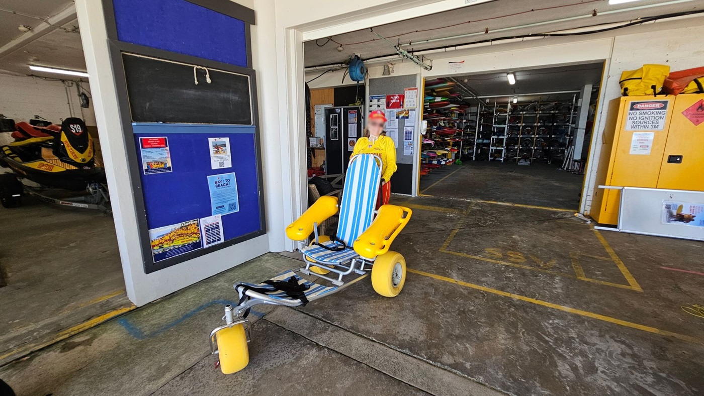 A garage-like space with a large open entrance. On the left, there's a blue notice board with various posters. Inside, a yellow and black jet ski is visible. In the centre, a person in a yellow shirt stands behind a beach wheelchair with blue and white stripes and large yellow wheels. On the right, shelves filled with equipment are visible, along with a yellow cabinet displaying safety notices. The floor is concrete with some painted lines.