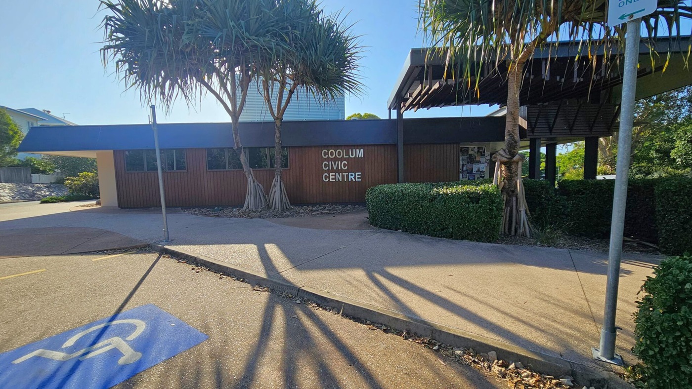 A street scene with a "No Through Road" sign on the left, surrounded by lush greenery and a large tree. The Coolum Civic Centre is in the centre, featuring a modern design with a slanted roof. To the right, more trees and a sign for Jack Morgan Park are visible. The pavement is a mix of concrete and asphalt, with shadows cast by the trees. The sun is partially visible, creating a bright spot in the sky.