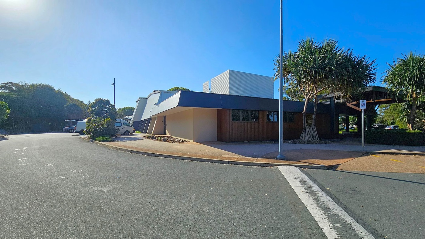 A modern building with angular architecture is on the right, featuring a dark overhang and a light-coloured facade. A tree with spiky leaves stands nearby, next to a parking sign. On the left, a few vehicles, including a white van, are parked along a curved road. The background is filled with dense greenery, and the sky is clear. The pavement is light-coloured, contrasting with the dark road surface.