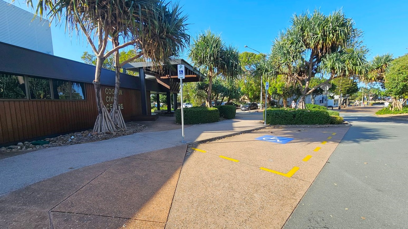 A paved area with a designated disabled parking space marked in blue and yellow lines is in the foreground. On the left, a building with a sign reading "Cooee Civic Centre" is partially visible, with large windows and a dark exterior. Several pandanus trees are scattered around, providing shade. In the background, a few cars are parked along a driveway lined with hedges and more trees. A clear blue sky is visible above.