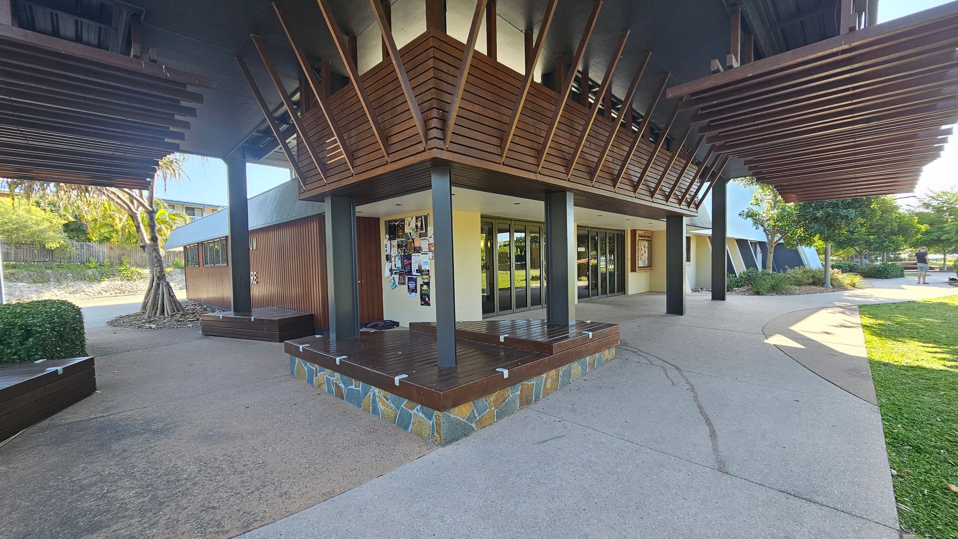 A modern building with a wooden exterior and angular design elements. The left side features a tree with exposed roots and a small hedge. The centre has a notice board with various posters and a stone base with wooden benches. The right side shows a pathway leading to a grassy area with trees and a person in the distance. The ceiling has wooden slats extending outward, creating a shaded area. The floor is a mix of concrete and stone.