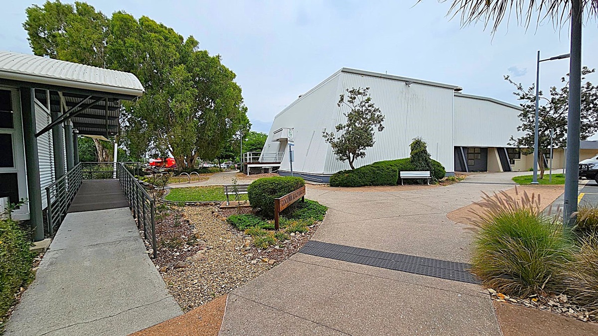 A modern building with a curved roof, identified as Coolum Beach Library, is in the centre. It has large windows and a ramp leading to the entrance. A signpost in the foreground indicates dogs must be on a leash. The surrounding area features well-maintained greenery and trees on both sides. The path in front is paved, with a small garden bed and a bicycle rack to the right.