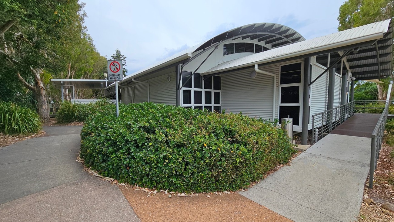 A modern building with a curved metal roof and large windows is on the right. A ramp leads up to the entrance. In the foreground, there is a dense green hedge. On the left, a path curves around the hedge, with a "Skateboards Prohibited" sign visible. Tall trees with thick trunks and green foliage are in the background, providing shade. The ground is a mix of concrete and tan-coloured paving.