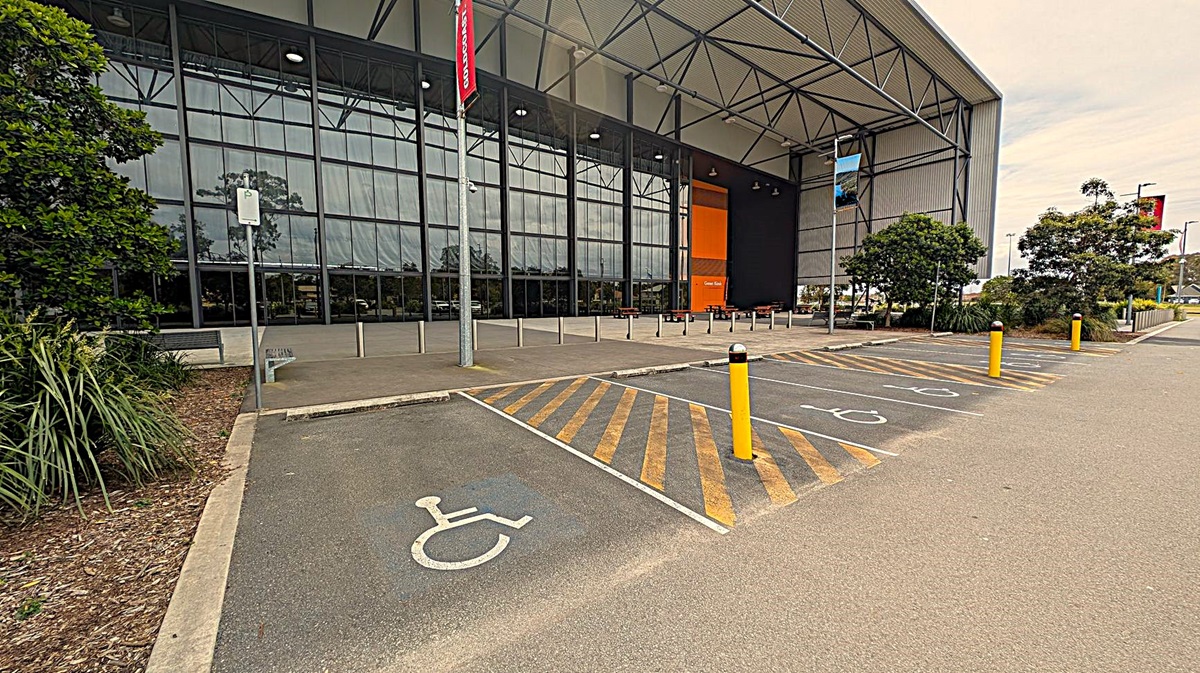 A large building with a high, angular roof and a facade made of glass and metal. In the foreground, there is a car park with several vehicles, including a blue utility vehicle on the left and a black pickup truck on the right. The entrance is flanked by trees and landscaped greenery. The building has a modern industrial design with visible structural supports. The ground is paved, and there are parking spaces and a pedestrian pathway leading to the entrance.