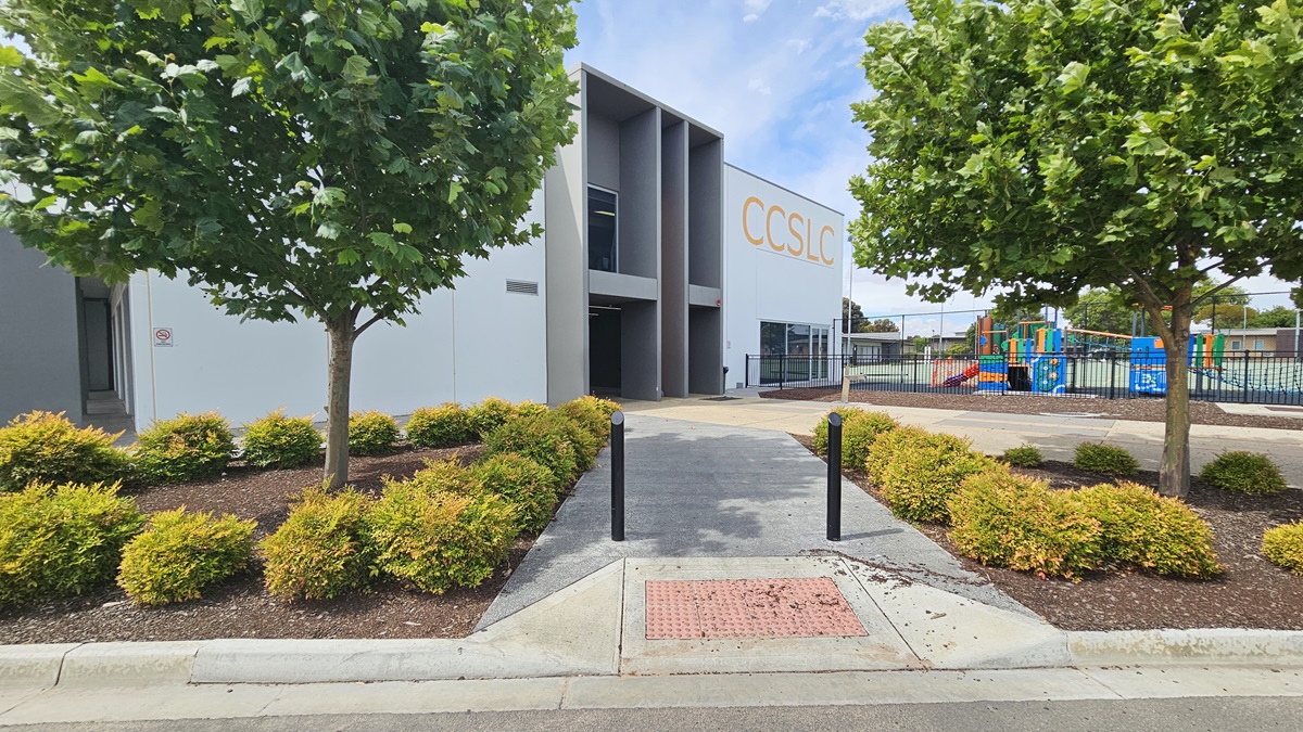 Copper Coast Sport and Leisure Centre is a large, white, and grey industrial building with a bold yellow-orange “CCSLC” sign on the left and front of the façade. The centre has glass entry doors and windows with white metal frames. Close to the left of the building are accessible car parking spaces, trees, and gardens with bushes maintained at a similar height. Right of the pavement leading to the entrance is a black, low, iron fence.