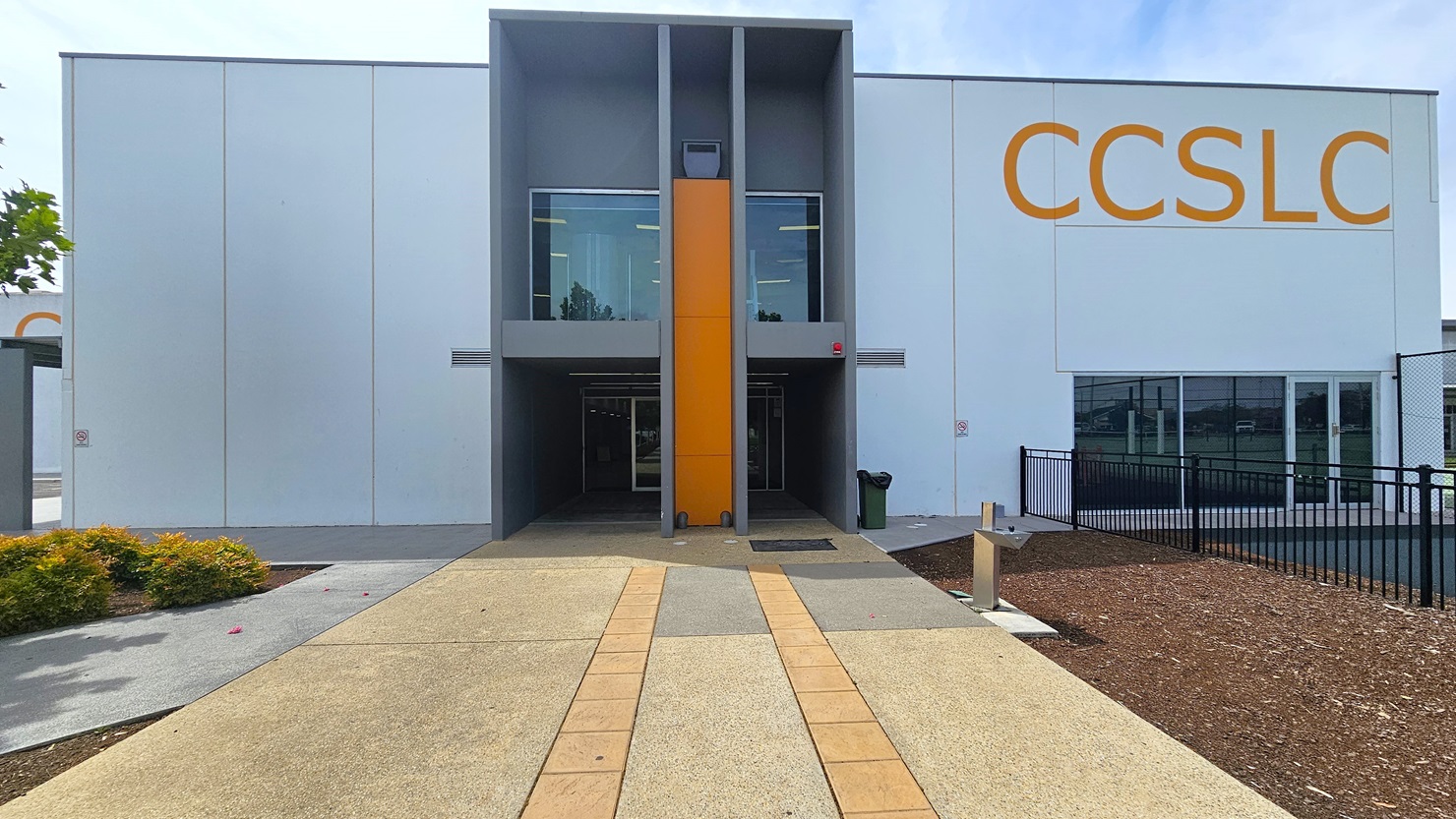 Central pathway leading to the entrance of the facility. The pathway is made of light brown natural stone tiles with two strips of light orange tiles. On both the left and right walls of the front façade are “No Smoking” signs. The entryway is divided by a central wall of orange tiles. The 2nd floor has glass windows with silver metal frames. There is a green garbage bin with a black bag on the right path leading to the children’s playground and tennis courts.