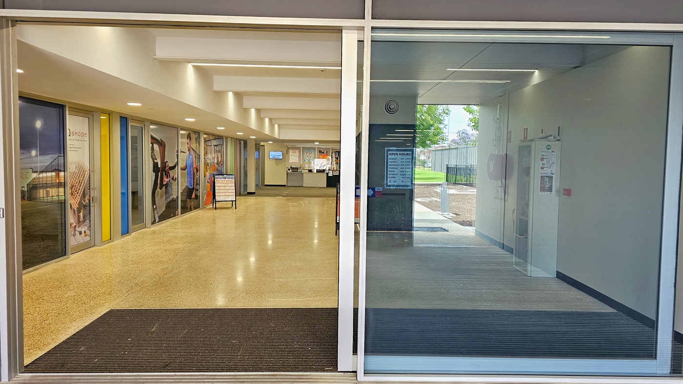 Automatic glass sliding doors with white metal frames at Copper Coast Sport and Leisure Centre. The entrance door is half open and shows a grey floor section right after the doors. Inside the centre are a light brown natural stone floor, a white ceiling, and several displays of adverts on the left glass wall of sports therapy clinicians' offices. The left ceiling has a drop ceiling type with LED COB lights, while in the middle space are rectangular blocks with fluorescents.