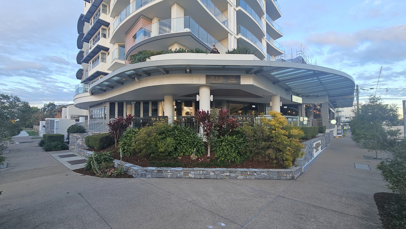 A multi-storey building with curved balconies on the left, featuring a ground-floor café with outdoor seating. The right side has a pathway leading to the entrance, bordered by landscaped greenery. In the middle, there are large windows and a glass canopy extending from the building. The foreground has a concrete pavement with a stone planter filled with various plants and shrubs.
