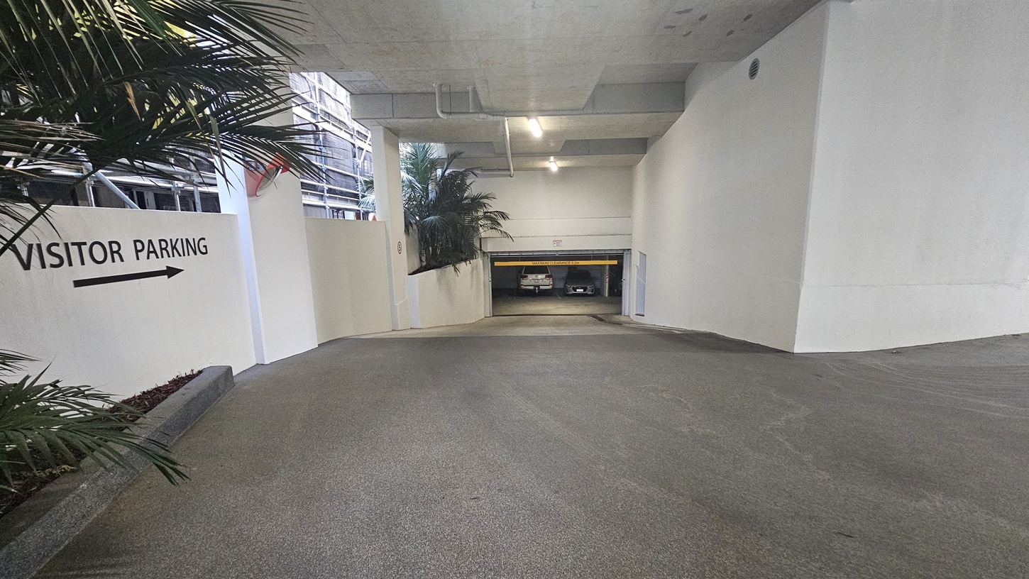 On the left, there is a white wall with a "Visitor Parking" sign and an arrow pointing right, next to a planter with tall palm plants. The right side has a plain white wall. In the middle, there is a sloped driveway leading down to an underground parking area with a yellow height restriction bar. The background includes parked cars and more palm plants. The floor is smooth concrete, and the ceiling is also concrete with exposed pipes and recessed lighting.