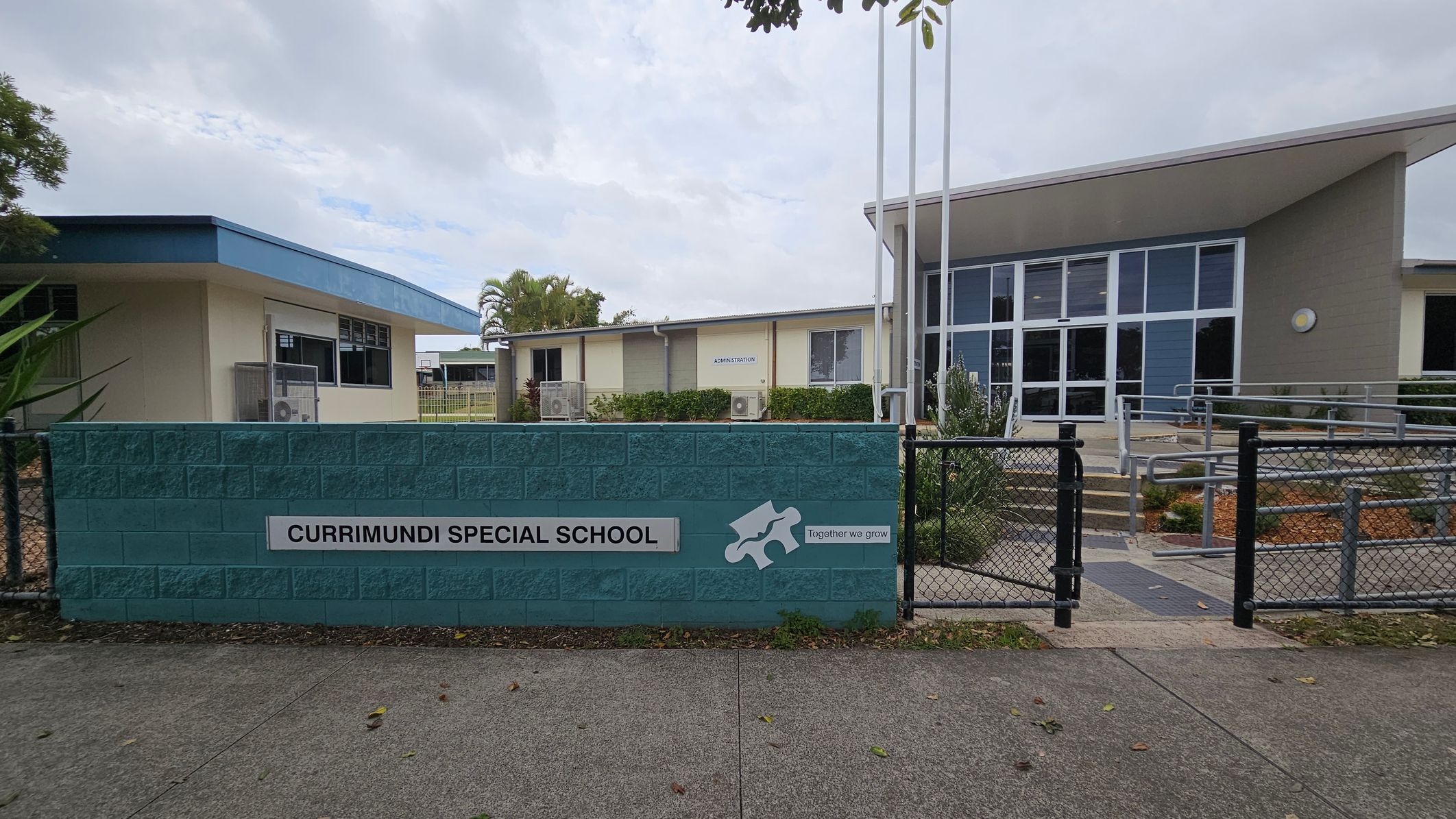 A school entrance with a blue brick wall in the foreground displaying the sign "Currimundi Special School" and the phrase "Together we grow" alongside a puzzle piece graphic. Behind the wall, there are two buildings. The left building has a light green exterior with a blue roof, while the right building features large glass windows and a sloped roof. A pathway with railings leads to the entrance. The ground is paved, and there are some plants and trees visible.