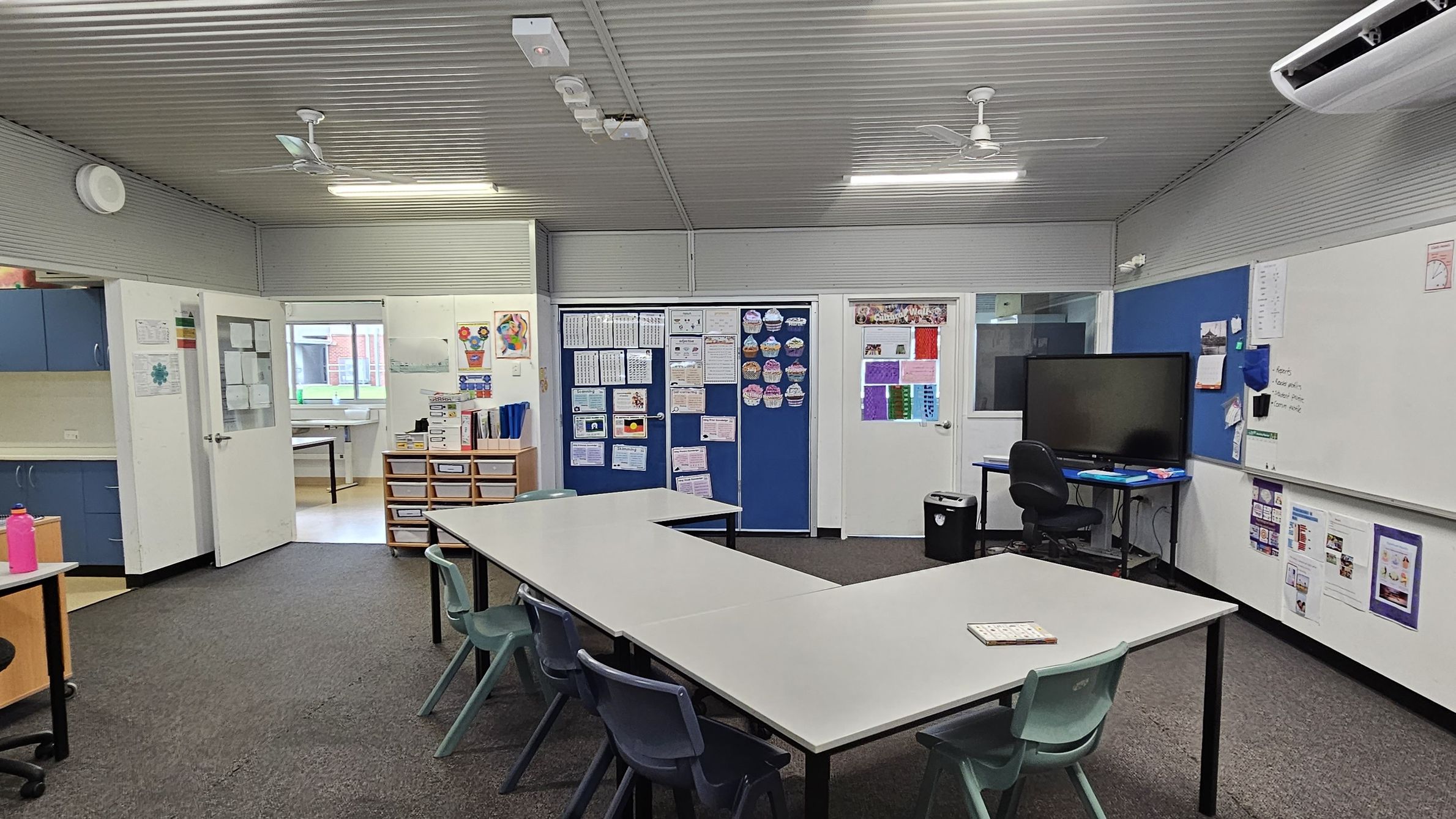 A classroom with a U-shaped arrangement of white tables and green chairs in the centre. On the right, there's a large whiteboard and a blue noticeboard with a flat-screen monitor on a desk. The back wall features blue and white doors with educational posters. On the left, blue cabinets and a pink water bottle are visible. The floor is carpeted, and the ceiling is corrugated with fluorescent lights and a ceiling fan. An air conditioning unit is mounted on the right wall.