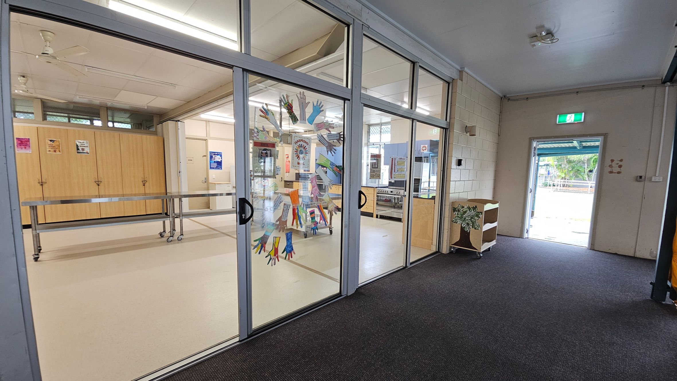 A room with glass sliding doors decorated with colourful handprints. Inside, there are wooden cabinets and a metal table on wheels. The ceiling has fluorescent lights and a ceiling fan. The floor is carpeted in the foreground and tiled inside the room. To the right, an open door leads outside, with a small shelf nearby. Posters are visible on the walls and cabinets.