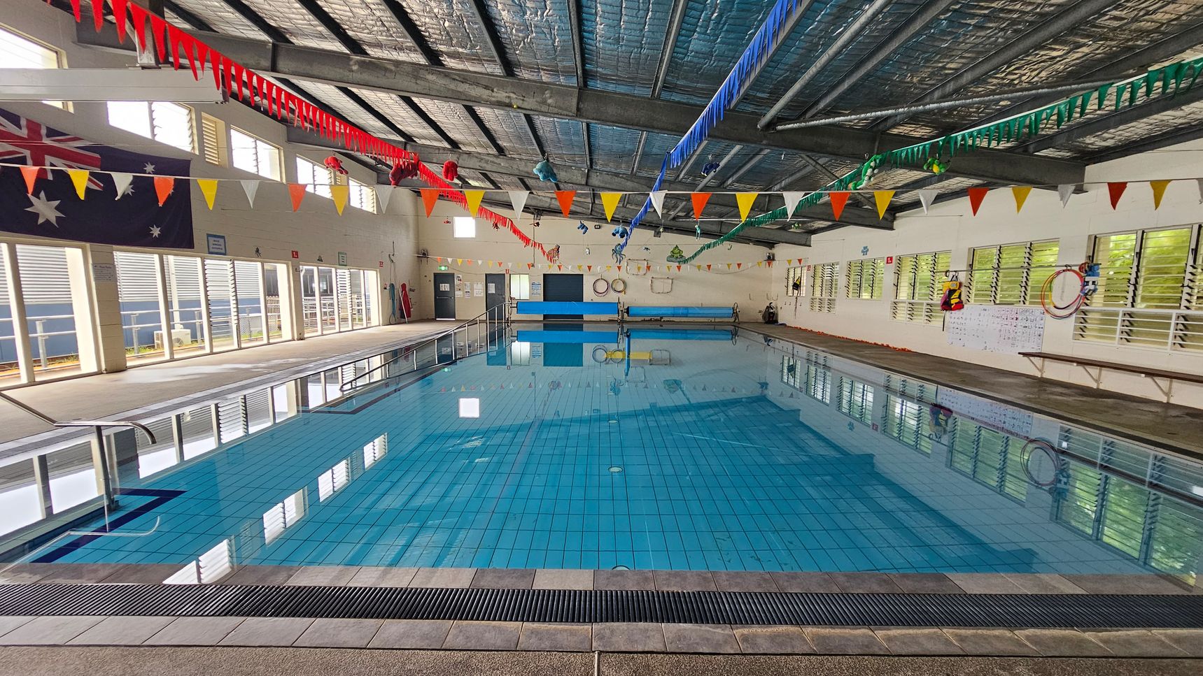 Indoor swimming pool with clear blue water, surrounded by a tiled deck. The ceiling is metal with exposed beams, adorned with colourful triangular bunting. On the left, large windows with louvres and an Australian flag. On the right, more windows and a wall with life rings and a notice board. The pool has a metal handrail on the left side. The floor is tiled, and the ceiling has insulation visible.