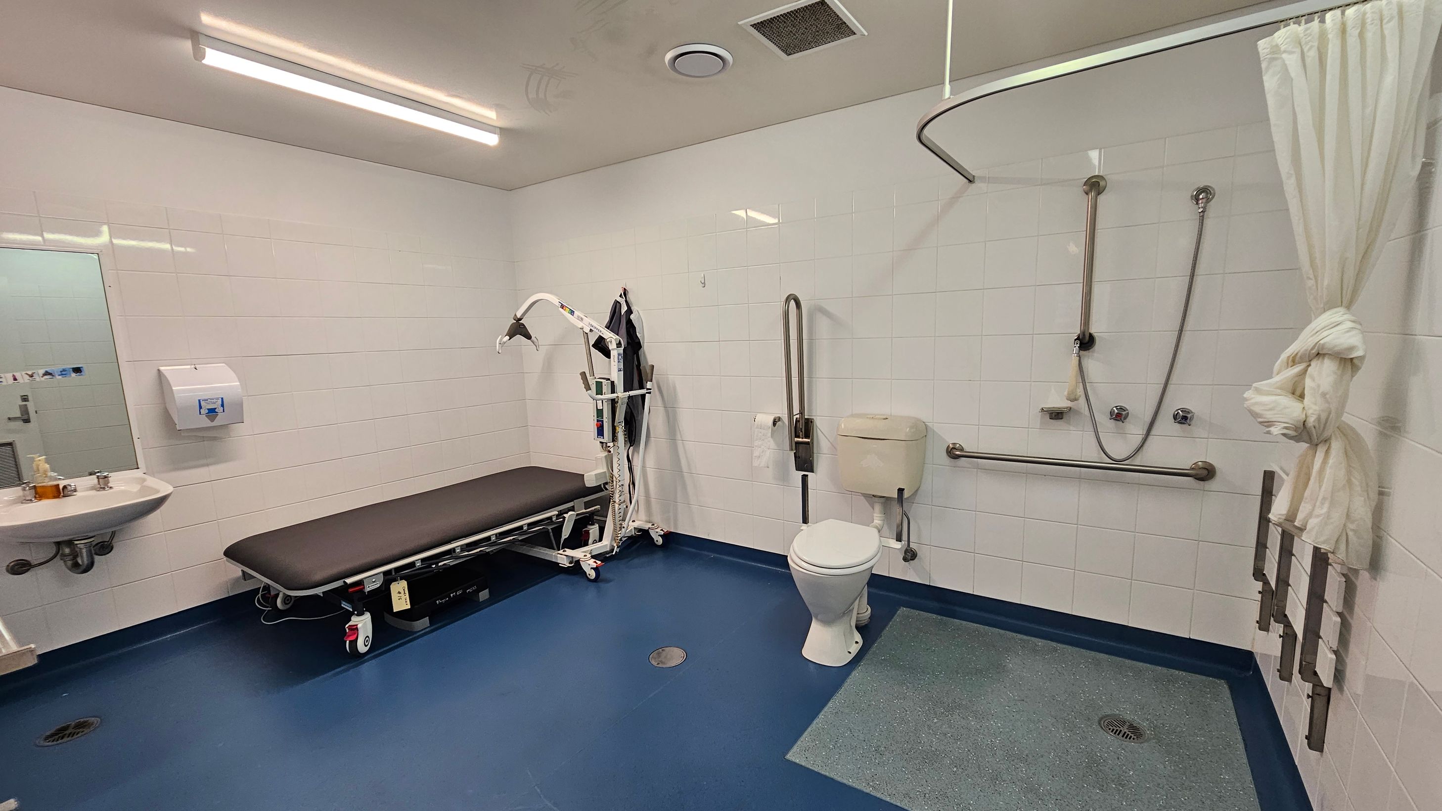 A spacious, accessible bathroom with white tiled walls and a blue floor. On the left, there's a sink with a mirror above it and a paper towel dispenser. A black cushioned bench with a hoist is positioned nearby. On the right, a toilet with grab bars is adjacent to a shower area with a curtain tied to the side. The ceiling has fluorescent lighting and ventilation fixtures.