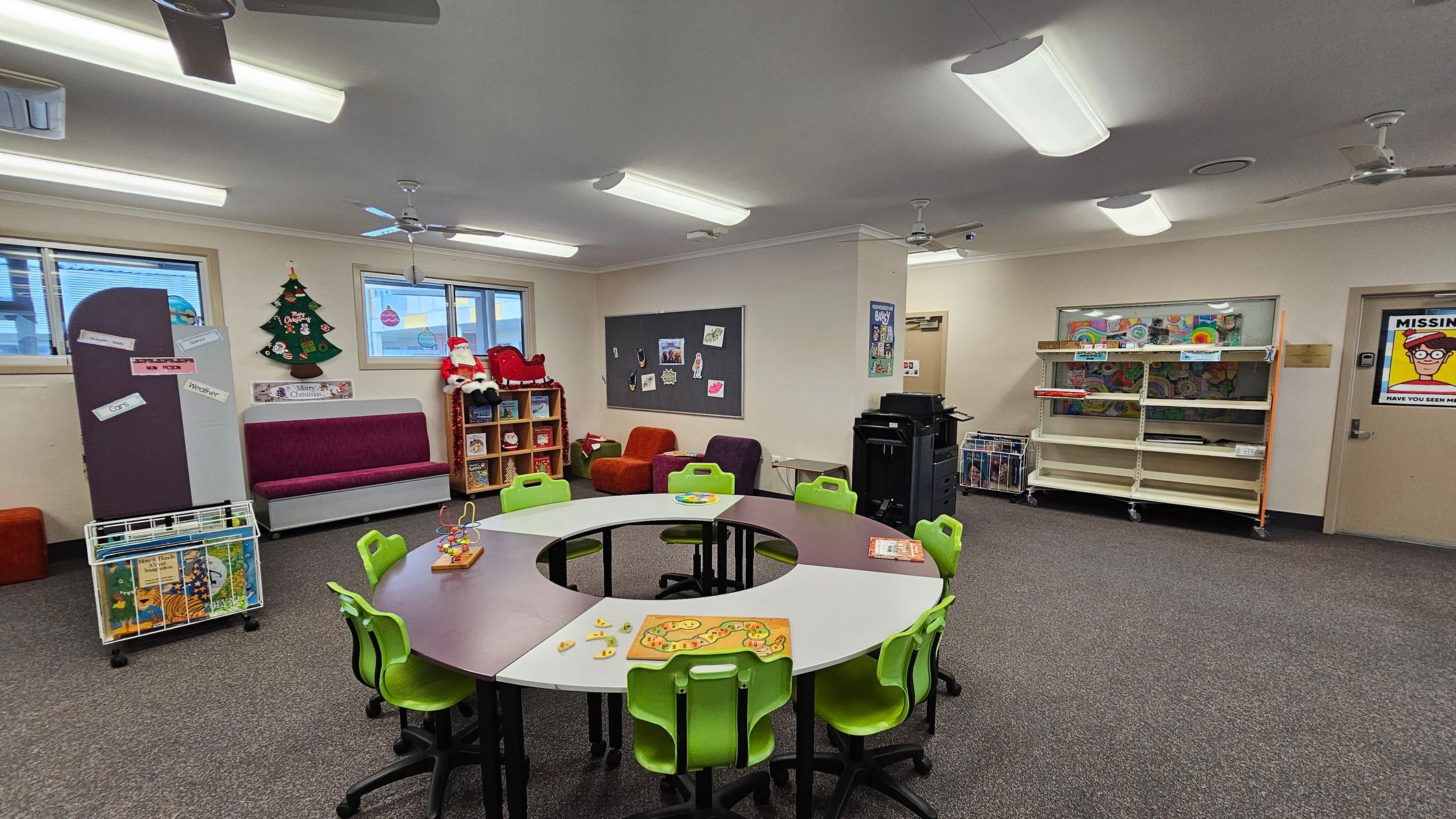 A classroom with a circular table in the centre surrounded by green chairs. On the table are educational toys and games. To the left, a purple bench sits beneath a window, with a Christmas tree decoration and a Santa figure on a shelf. The right side has a large bulletin board with artwork and a printer nearby. Shelves with books and a "Missing" poster are on the far right wall. The ceiling has fluorescent lights and fans, and the floor is carpeted.