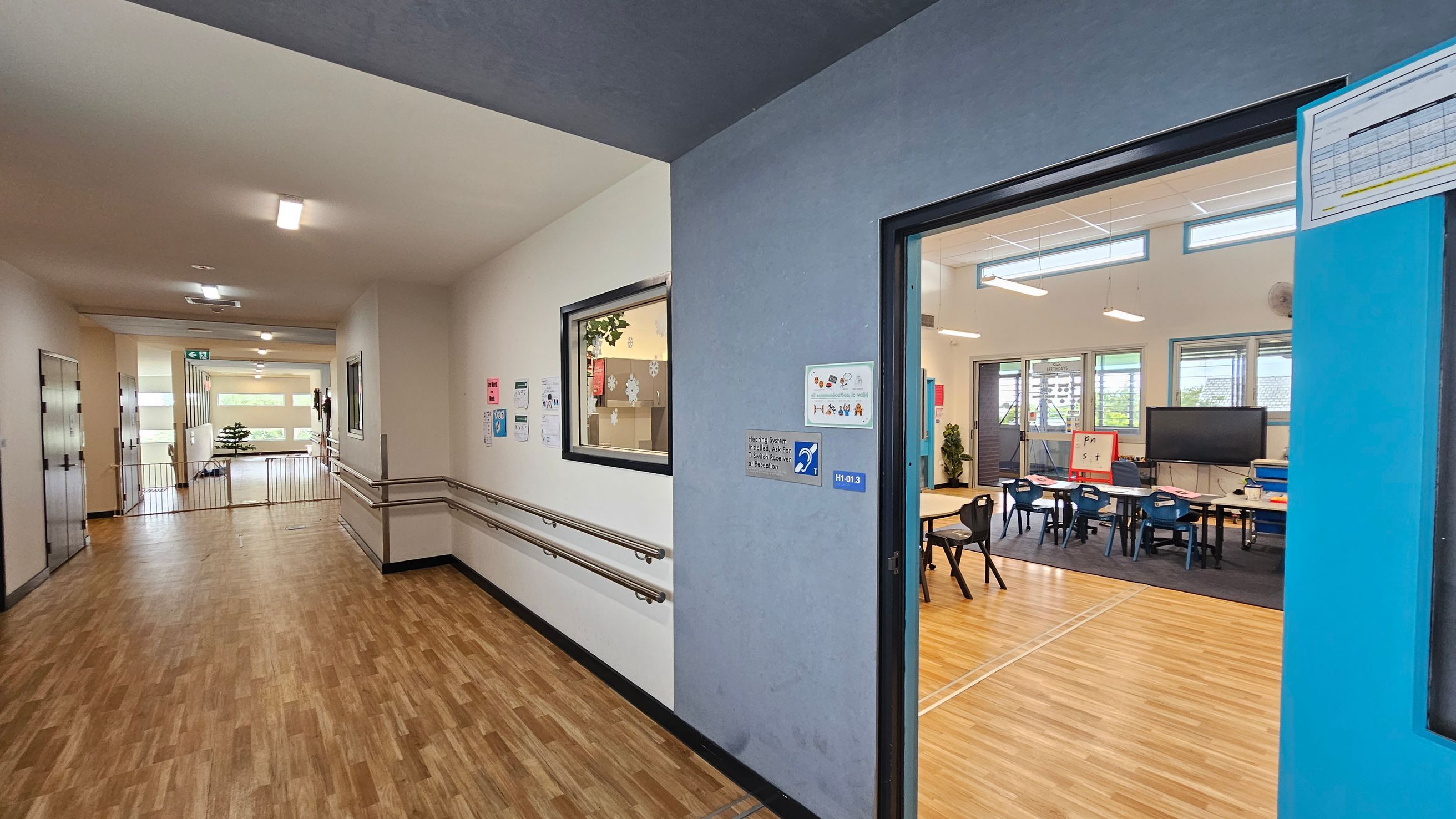 A school corridor with wooden flooring and white walls. On the left, there are handrails along the wall and a window with a view into a room. The corridor leads to a bright area with large windows and a small plant. On the right, an open door reveals a classroom with tables, chairs, a large screen, and a whiteboard. The classroom has light wooden flooring and several windows allowing natural light. Ceiling lights are visible in both the corridor and the